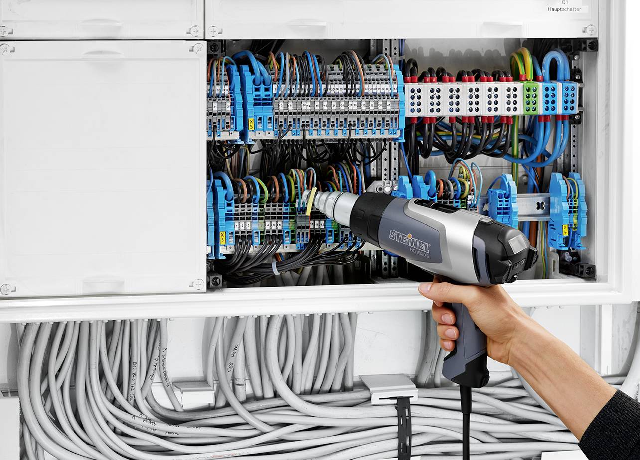 Person richtet mit einem Heißluftgebläse Kabel in einem offenen Schaltschrank. Verschiedene Kabel sind sorgfältig organisiert.