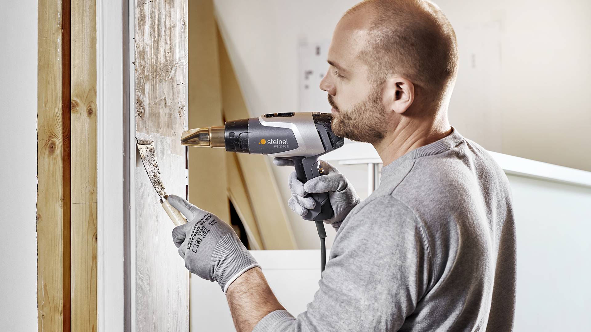 Mann entfernt Farbe von einem Fensterrahmen mit einem Heißluftgebläse und einem Spachtel in einem hell beleuchteten Raum.