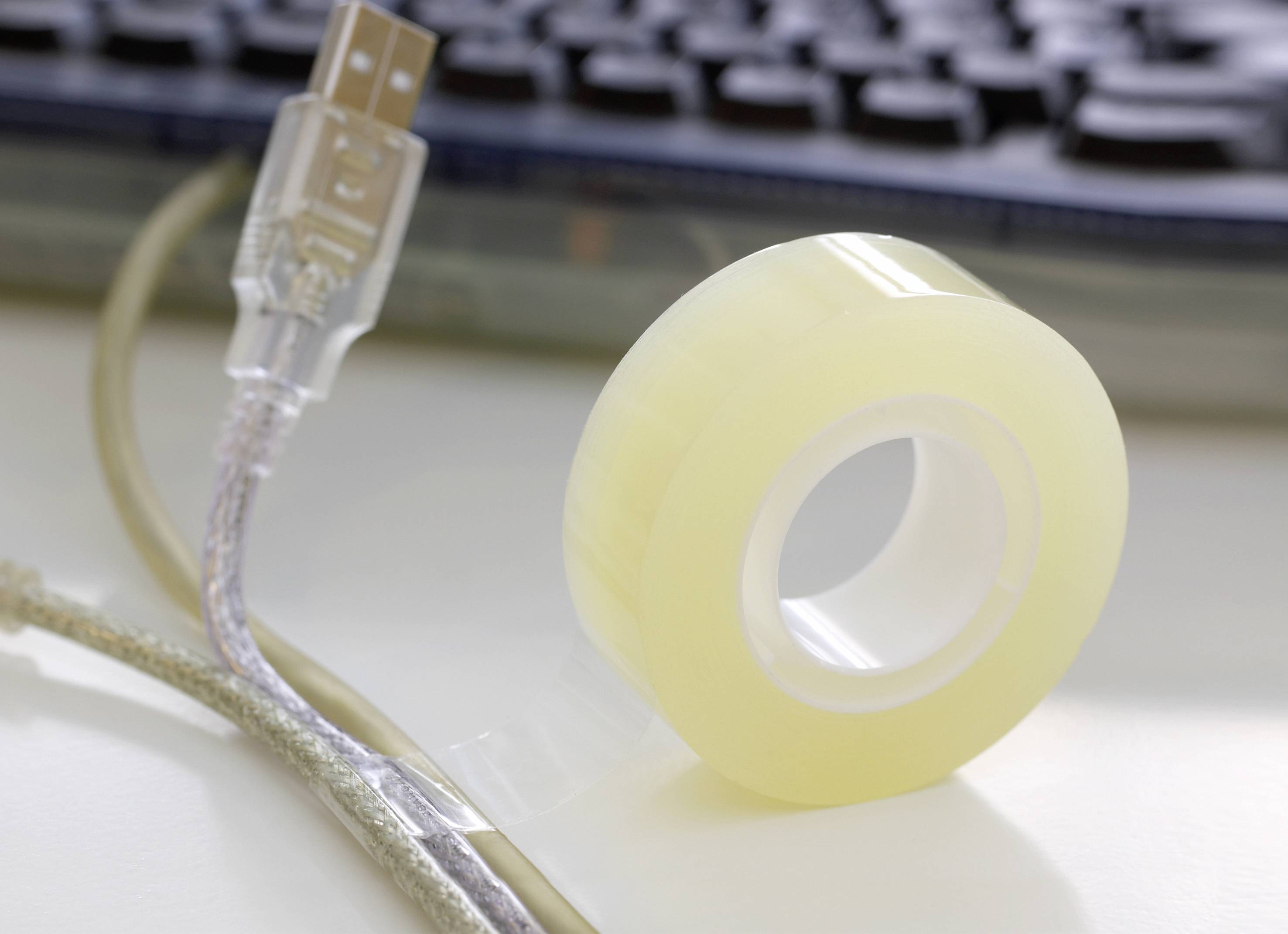 Klebebandrolle und USB-Kabel auf einem Tisch, teilweise verdecktes Computer-Keyboard im Hintergrund.