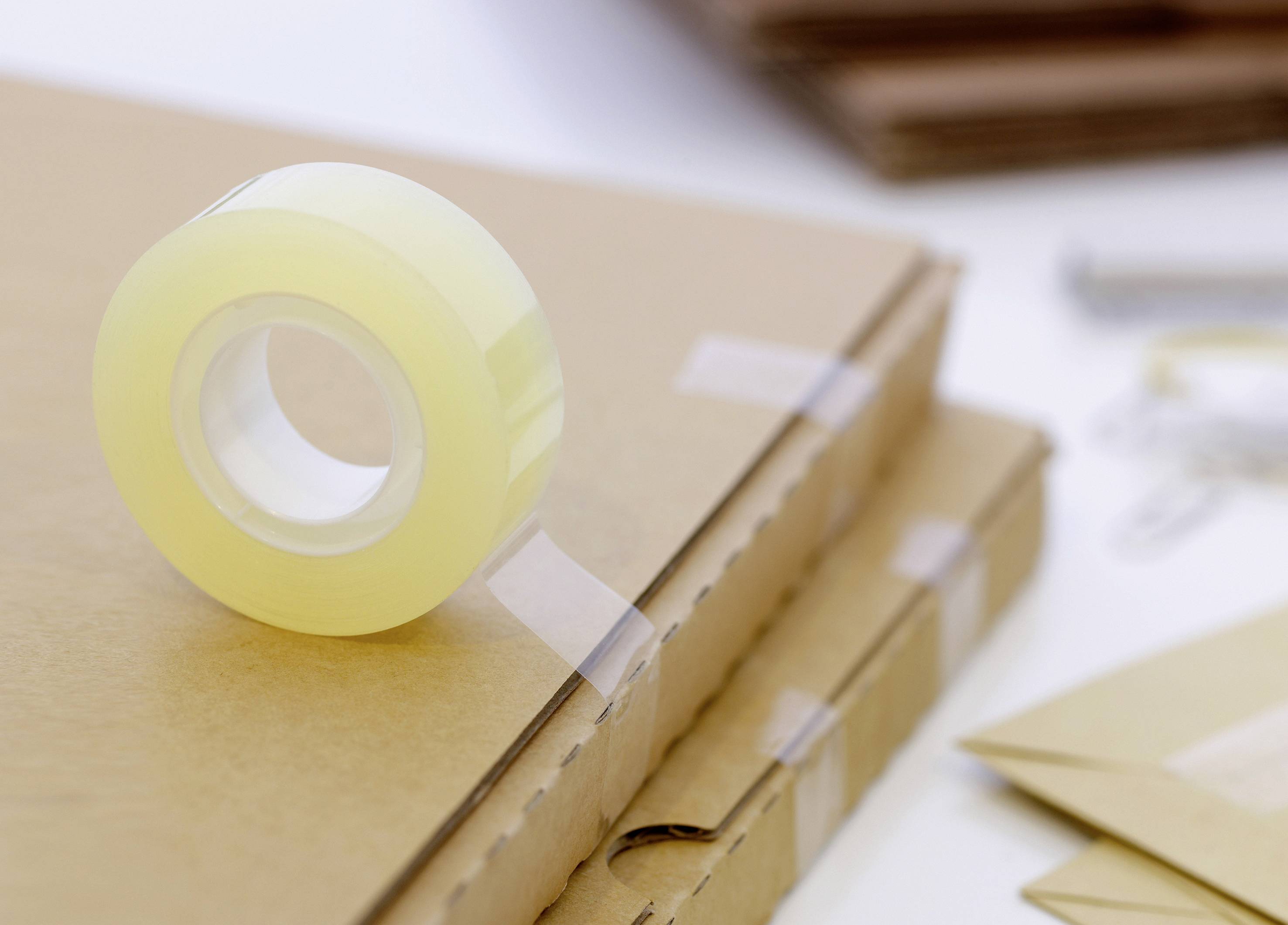 Eine Rolle Klebeband liegt auf mehreren gestapelten Kartons. Es gibt Büroklammern und Umschläge im Hintergrund.