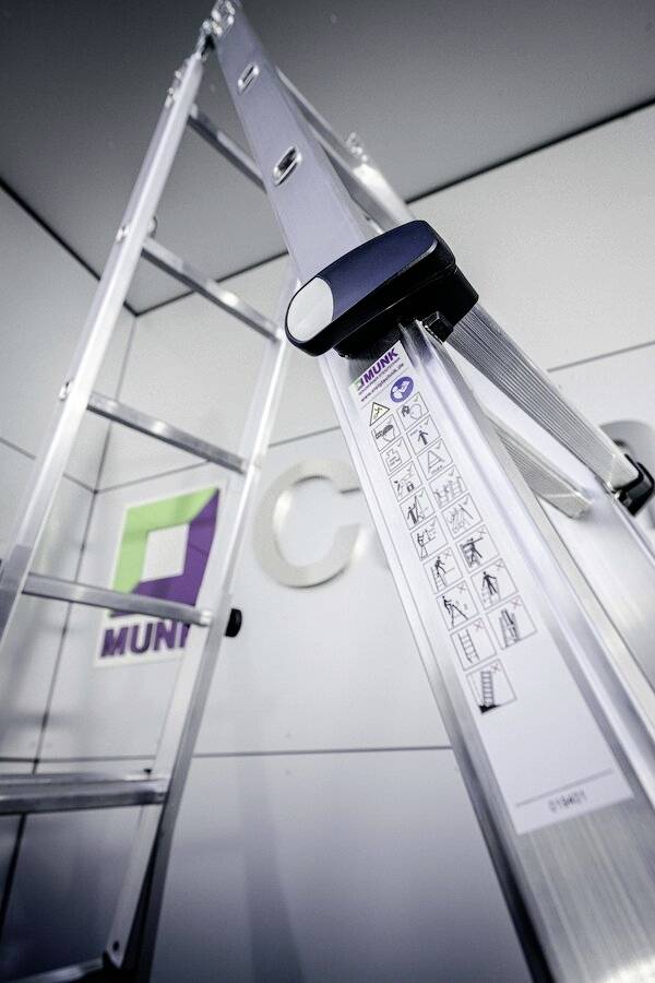 Aluminiumleiter gegen Wand gelehnt, mit eingezeichneten Sicherheitshinweisen. Firmenlogo im Hintergrund.