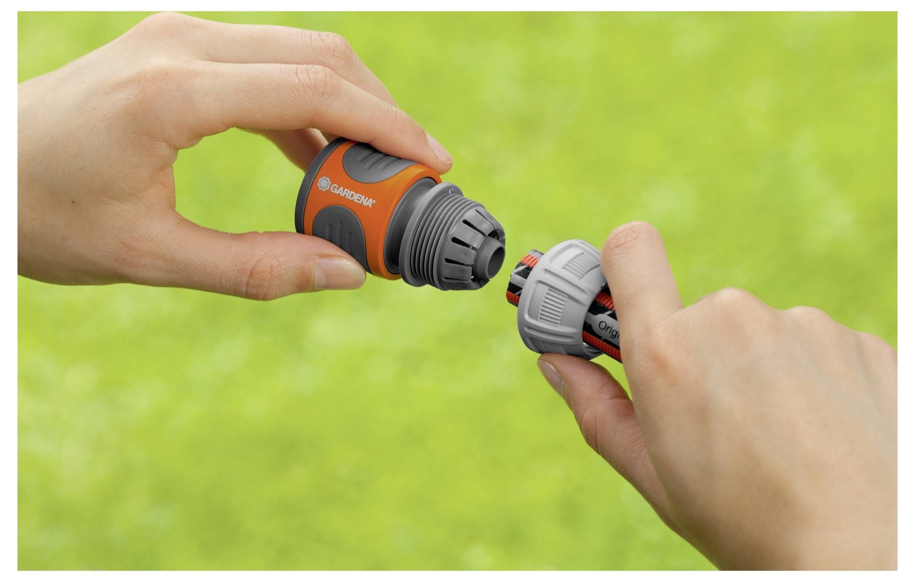 Hände verbinden einen Gartenschlauch mit einer orange-grauen Düse vor einem grünen Rasen im Hintergrund.