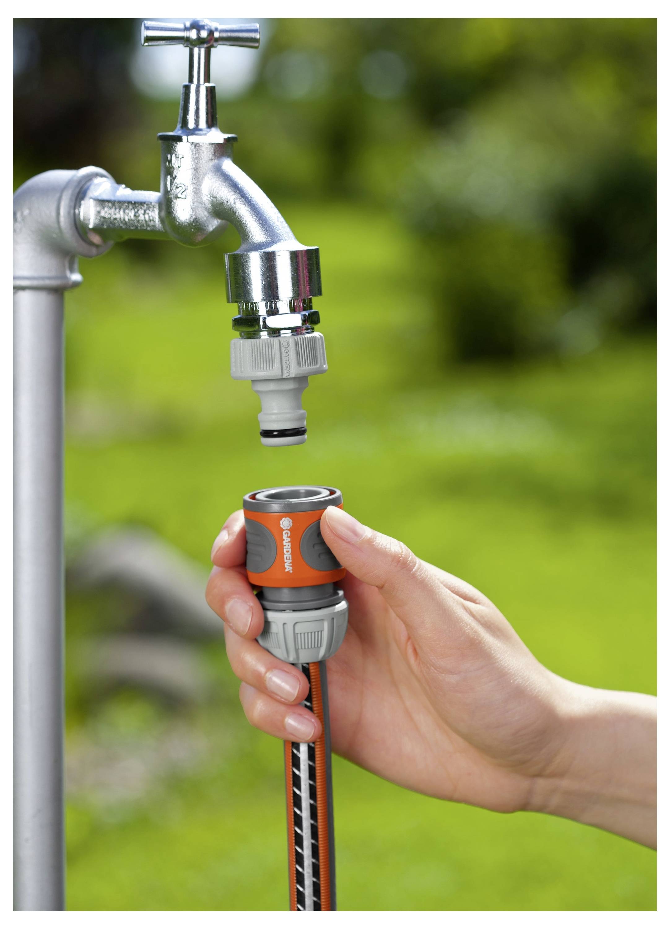 Eine Hand verbindet einen Gartenschlauch mit einem metallenen Außenwasserhahn mit Hilfe eines orange-grauen Adapters, mit einem unscharfen grünen Hintergrund.