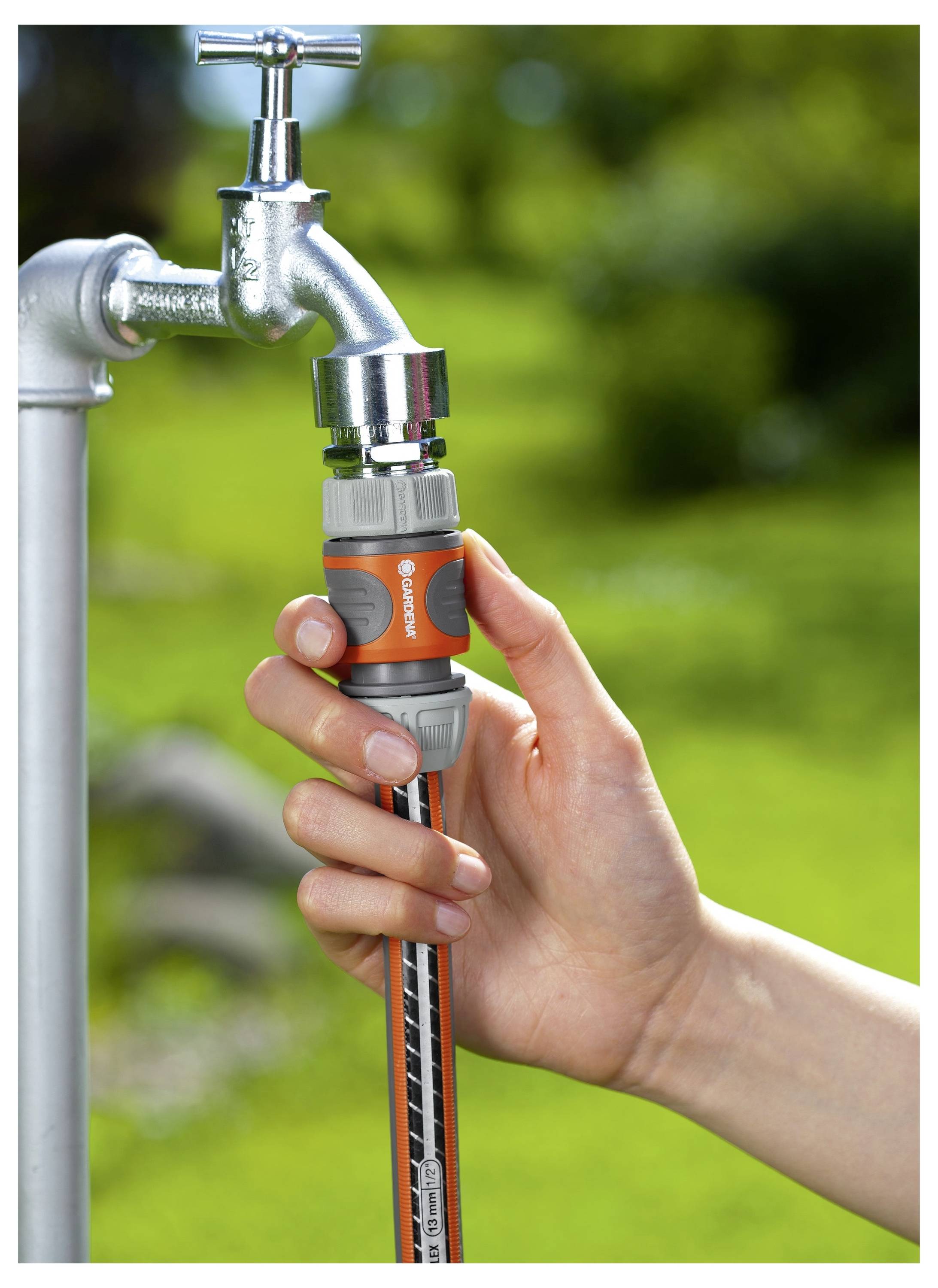 Eine Person verbindet einen orangefarbenen und grauen Schlauchaufsatz mit einem metallenen Außenwasserhahn. Der Hintergrund zeigt einen unscharfen grünen Garten.