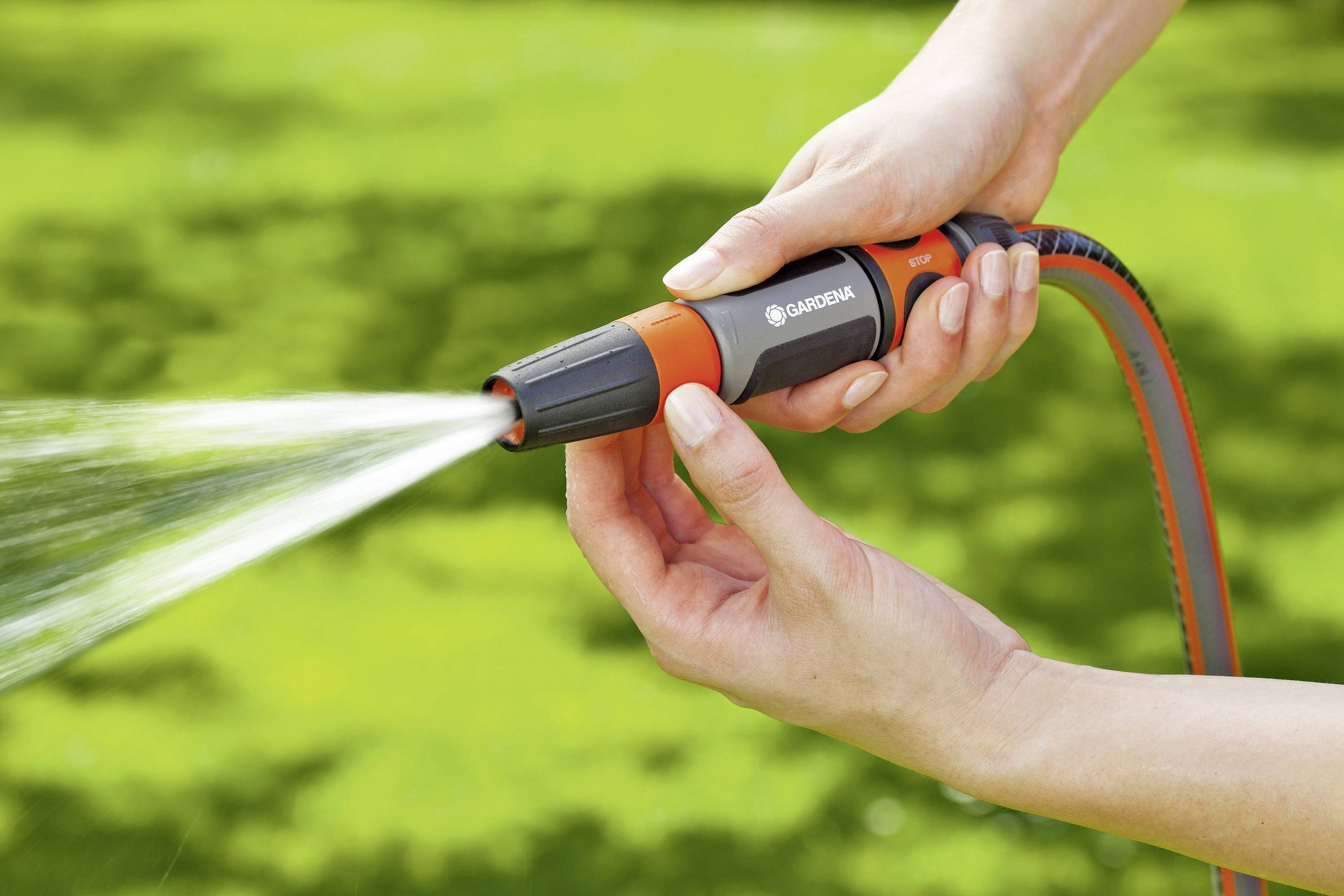 Person hält einen Gartenschlauch mit Sprühdüse, gießt Wasser in einen grünen Gartenhintergrund. Hände regulieren den Wasserstrahl.