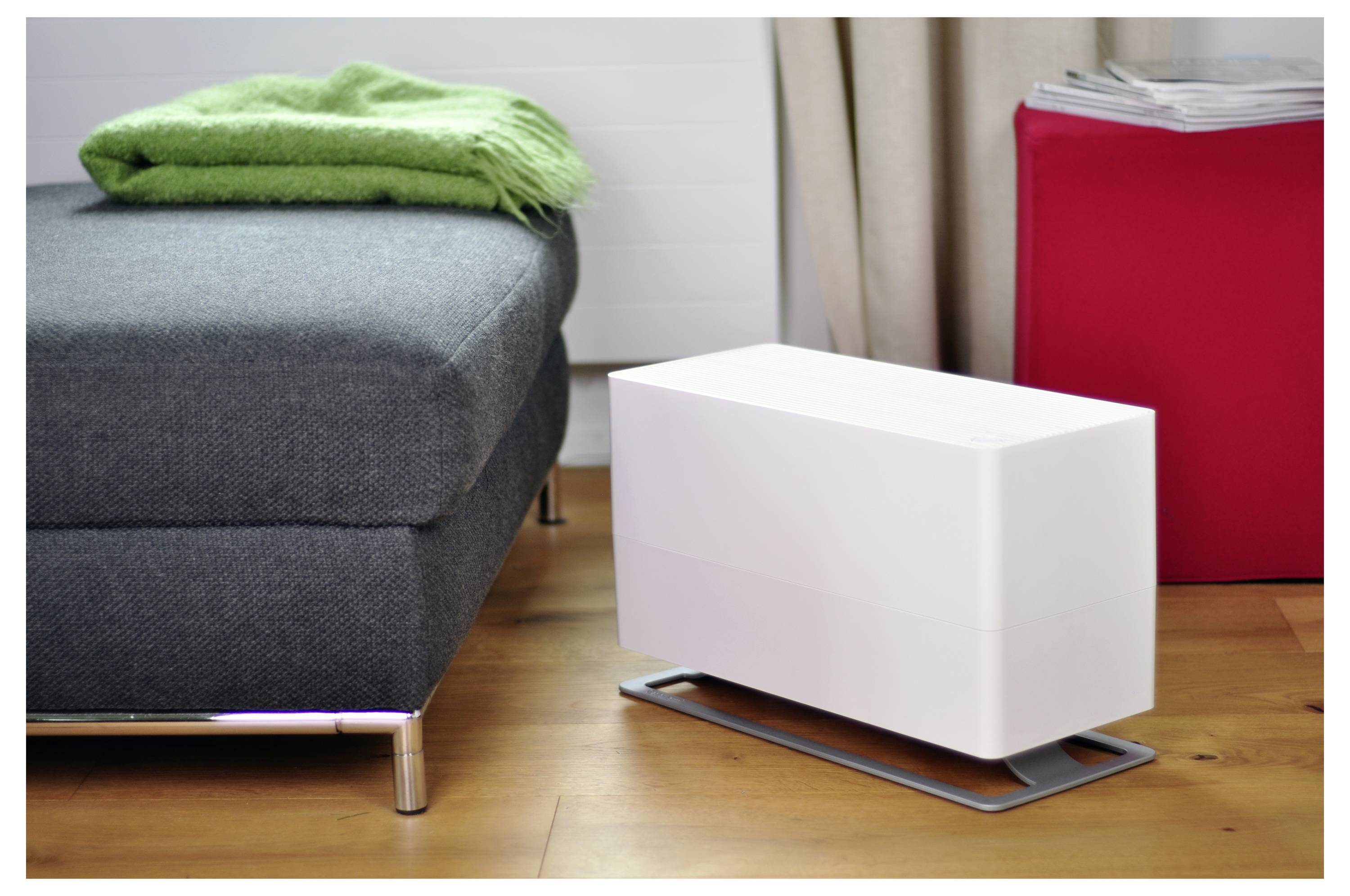 Weißer rechteckiger Luftreiniger oder Befeuchter auf dem Boden neben einem grauen Sofa mit grüner Decke und einem roten Schrank in einem Wohnzimmer.