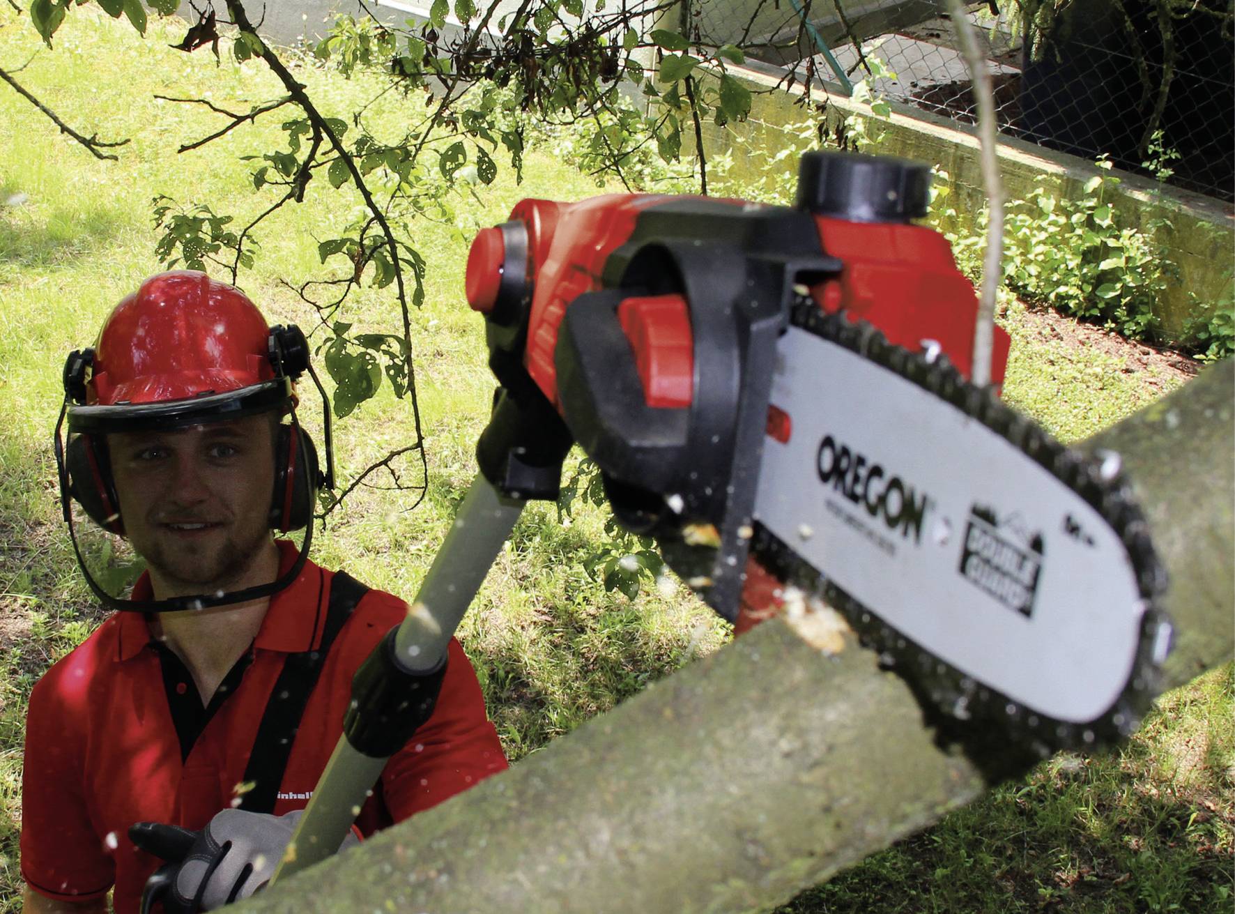 Eine Person trägt Schutzkleidung und verwendet eine elektrische Kettensäge mit Teleskopstiel, um Zweige von einem Baum abzusägen.