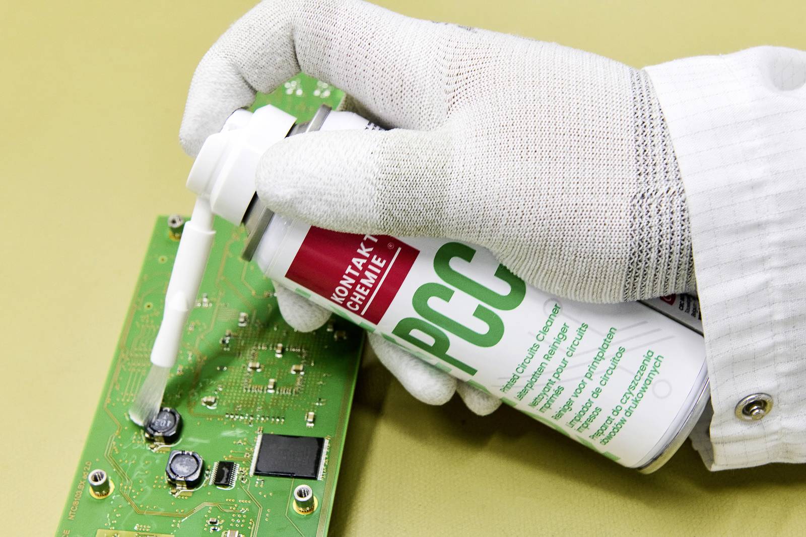 Eine behandschuhte Hand sprüht ein Reinigungsmittel auf eine grüne Leiterplatte, um elektronische Komponenten zu reinigen oder zu warten.