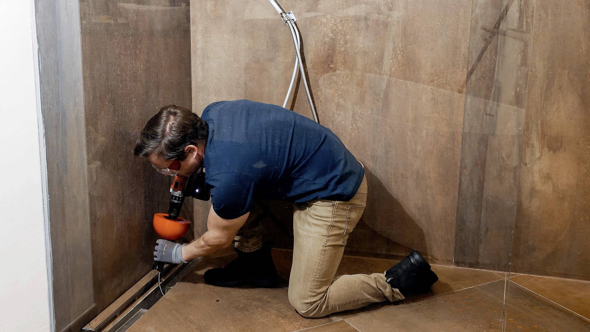 Ein Handwerker installiert eine Duschablaufrinne im Badezimmer. Er kniet auf dem Boden und arbeitet mit einem Werkzeug an der Abflussrinne.