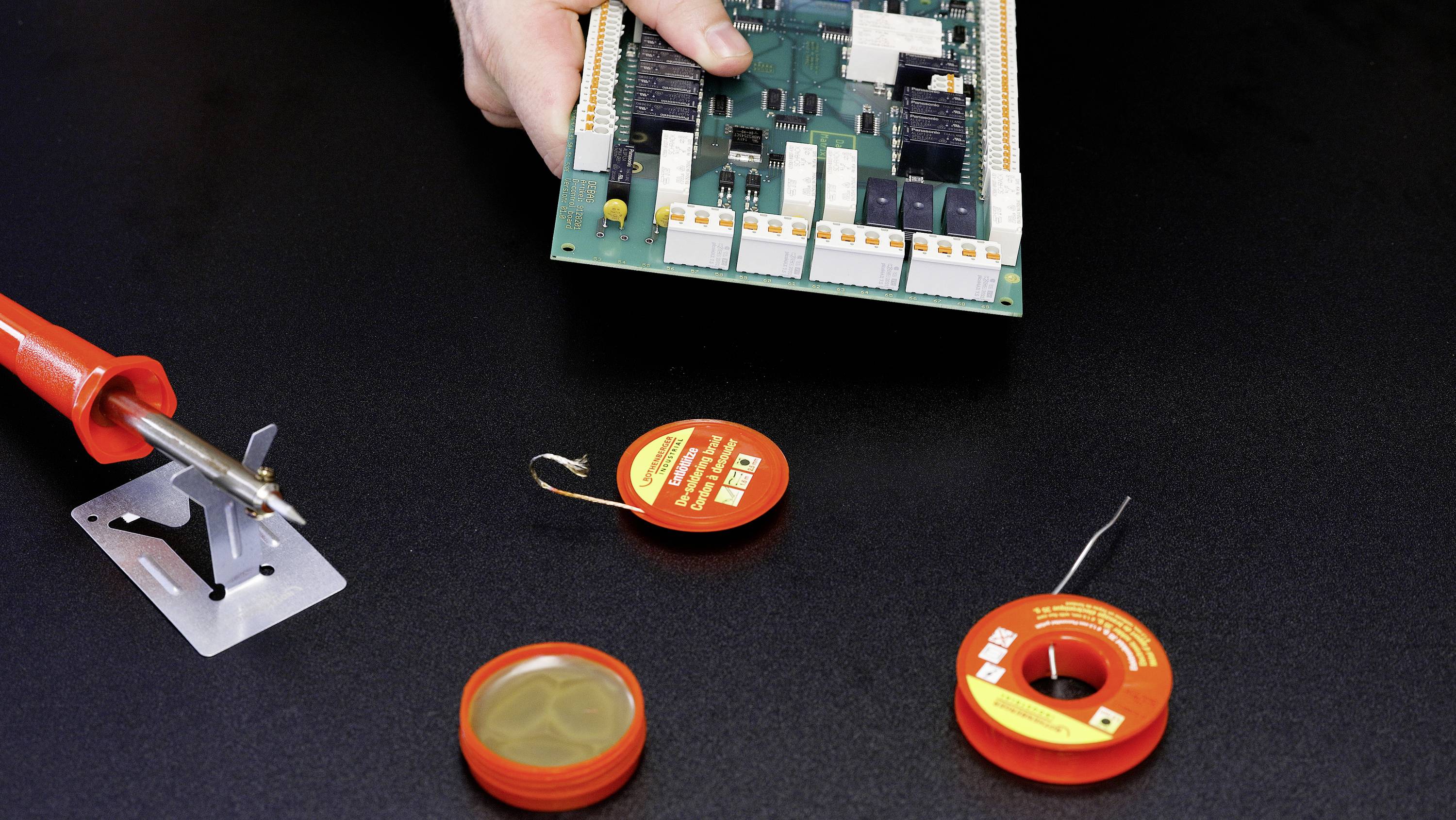 Eine Person hält eine elektronische Platine über einem schwarzen Tisch, der mit einem Lötkolben und Lötmaterialien ausgestattet ist.