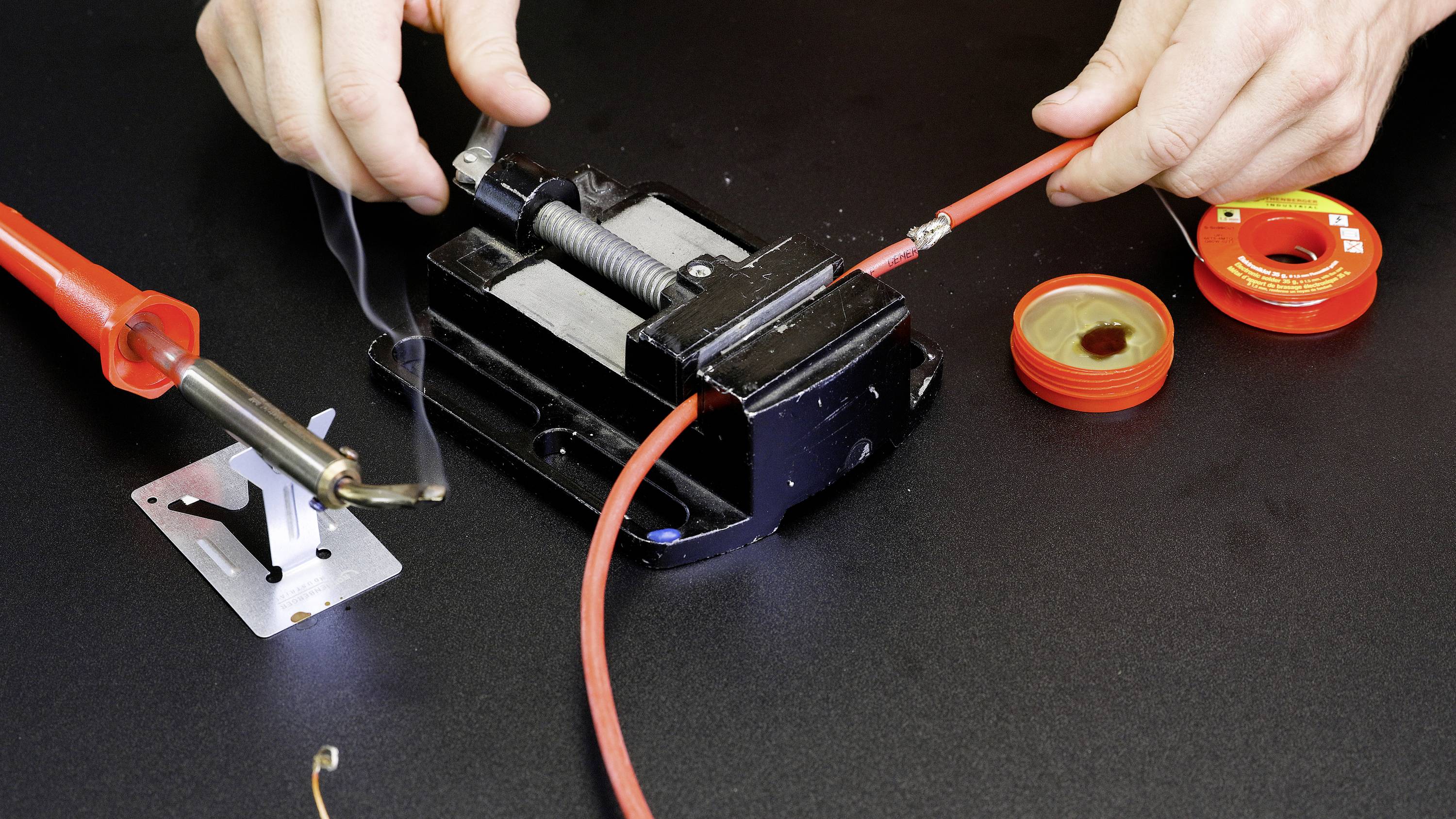 Person lötet ein rotes Kabel mit einem Lötkolben auf einer schwarzen Oberfläche; Rauch steigt vom Kabel auf, Werkzeug im Hintergrund.