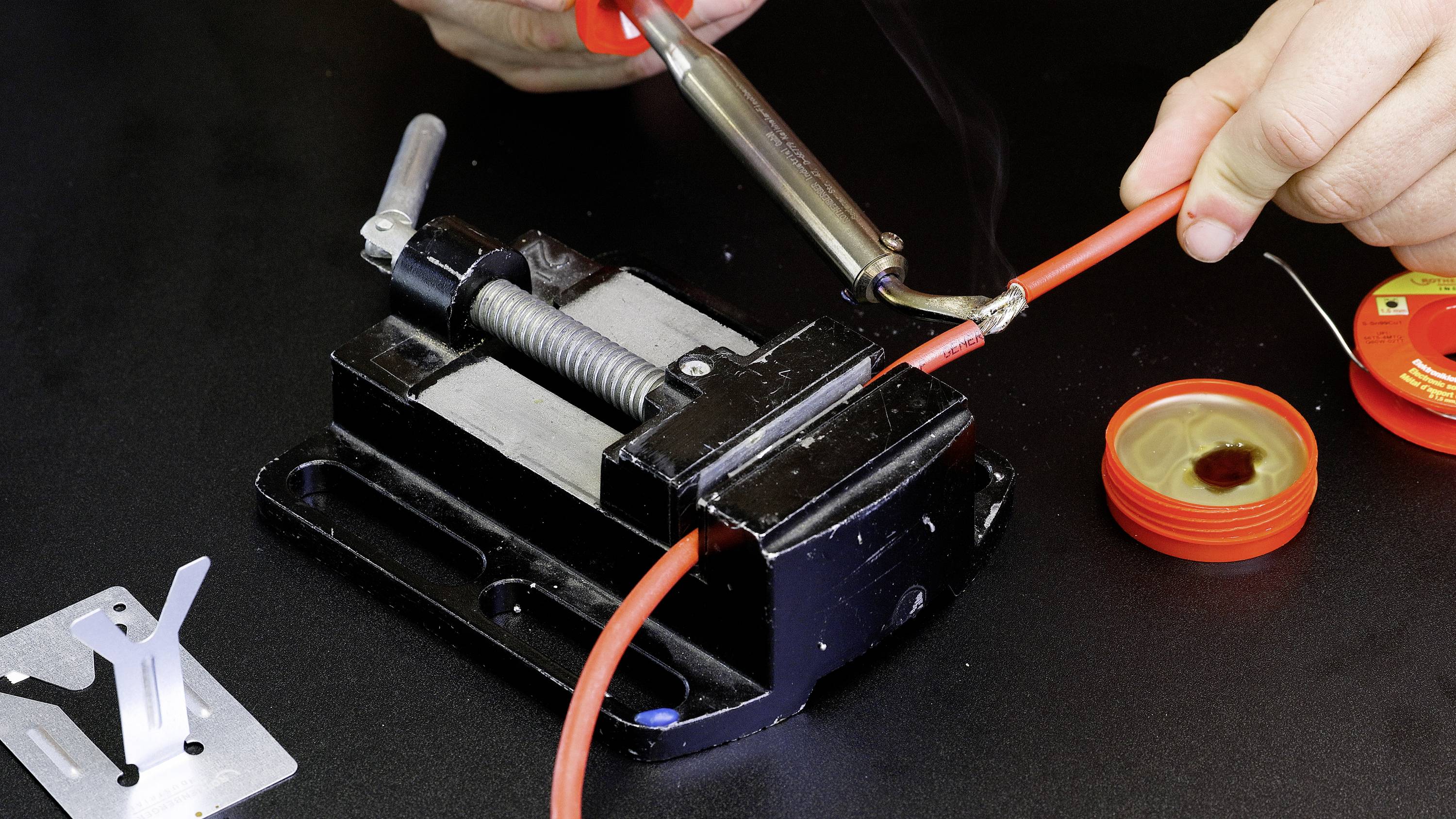 Eine Person lötet ein rotes Kabel, das in einem Schraubstock eingespannt ist. Daneben liegt Lötdraht und ein offener Lötmitteldeckel.