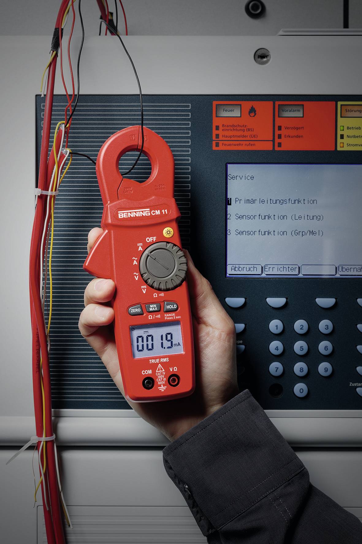 Eine Person hält ein rotes digitales Multimeter vor einem Schaltschrank und misst Spannung; Display zeigt '00.19'.