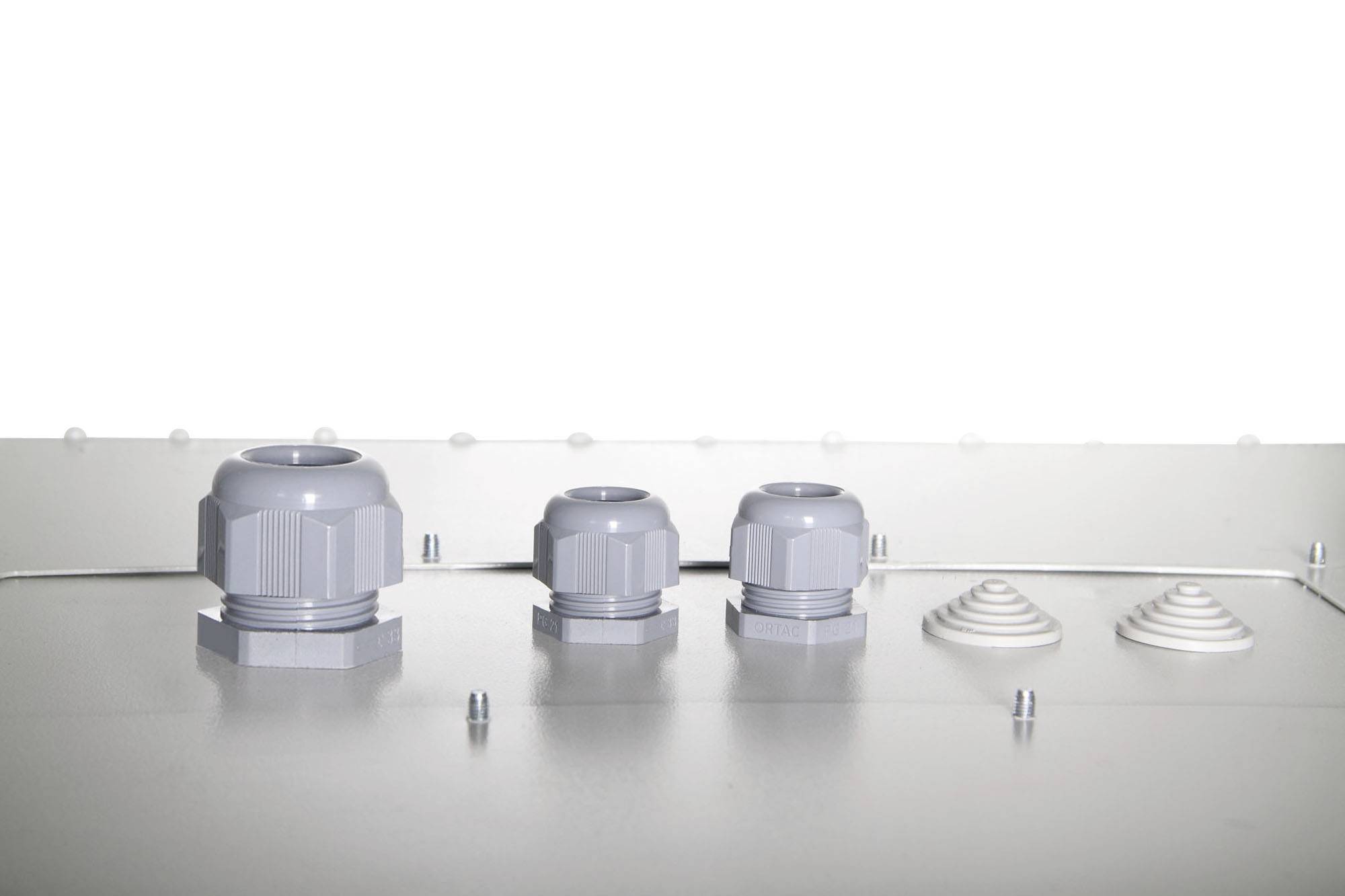 Drei graue Kabelverschraubungen unterschiedlicher Größe auf einer metallischen Oberfläche mit zwei weißen, kegelförmigen Objekten daneben.