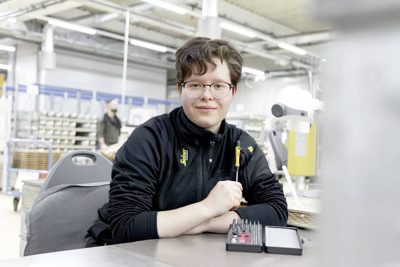 Eine lächelnde Person mit Brille sitzt in einer Werkstatt. Sie trägt einen schwarzen Pullover und hält einige Werkzeuge. Im Hintergrund sind Regale und eine weitere Person erkennbar.