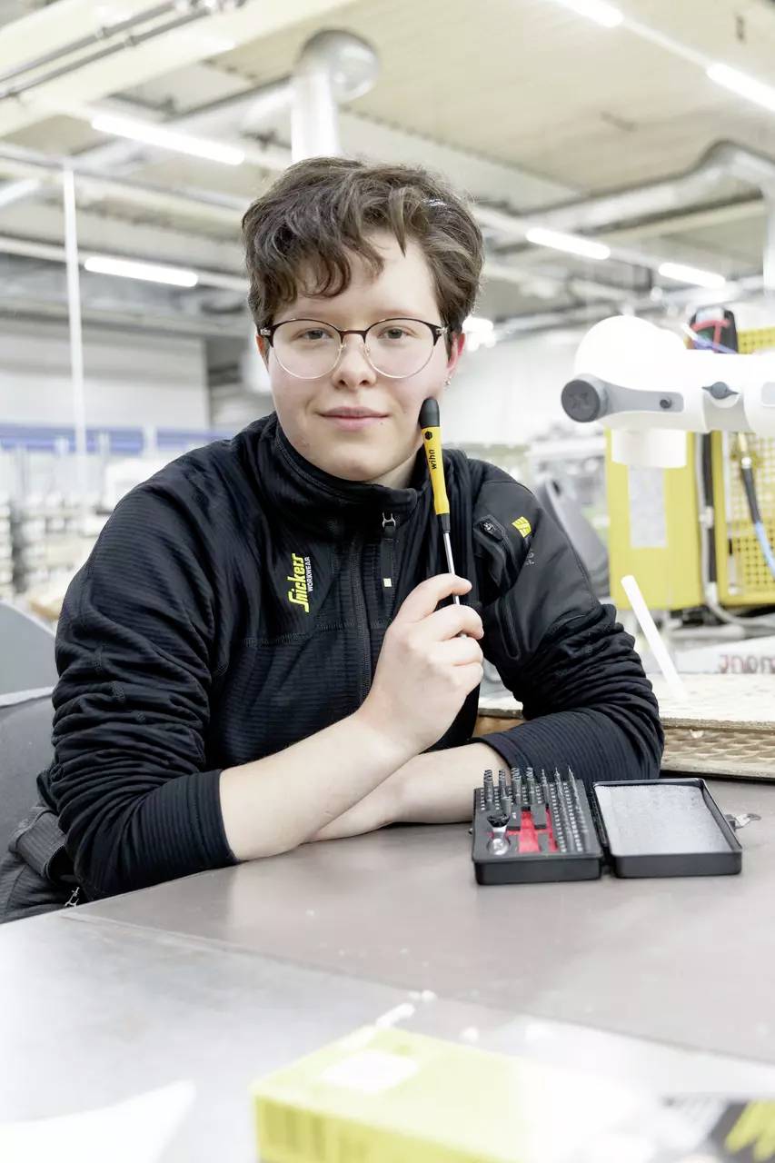 Eine Person in einer Werkstatt hält einen Stift und lächelt in die Kamera. Auf dem Tisch liegen Werkzeuge. Im Hintergrund sind industrielle Geräte.