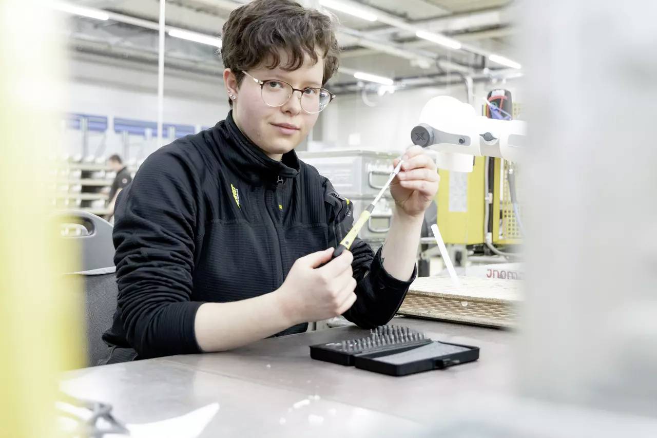 Eine Person arbeitet konzentriert in einer Fabrikumgebung und hält ein elektronisches Bauteil. Im Hintergrund sind Maschinen zu sehen.