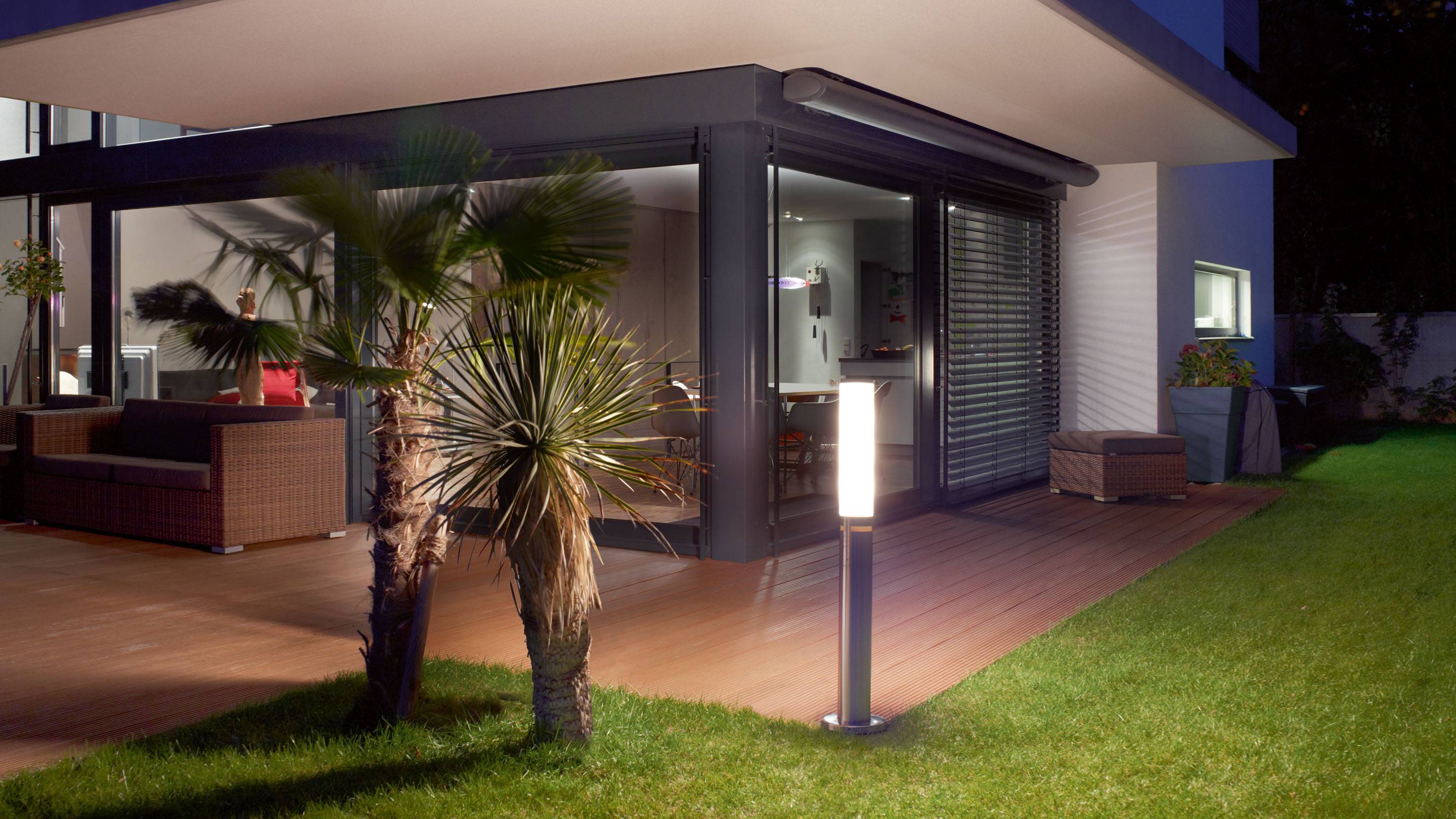 Ein modernes Haus mit einer Terrasse aus Holz, beleuchtet von Außenlampen. Pflanzen und Gartenmöbel schaffen eine gemütliche Atmosphäre.
