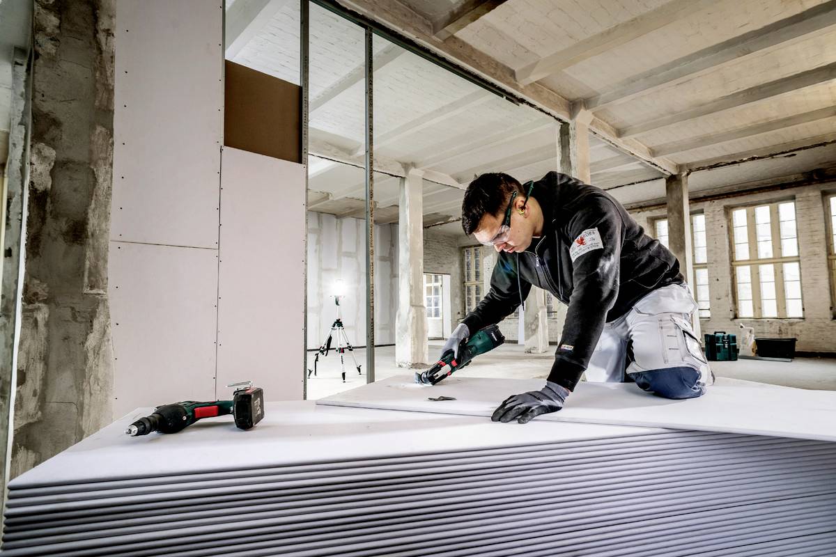 Ein Handwerker schneidet in einem Gebäudebauprojekt Trockenbauplatten. Er trägt Schutzbrille und arbeitet konzentriert mit einem Elektrowerkzeug.