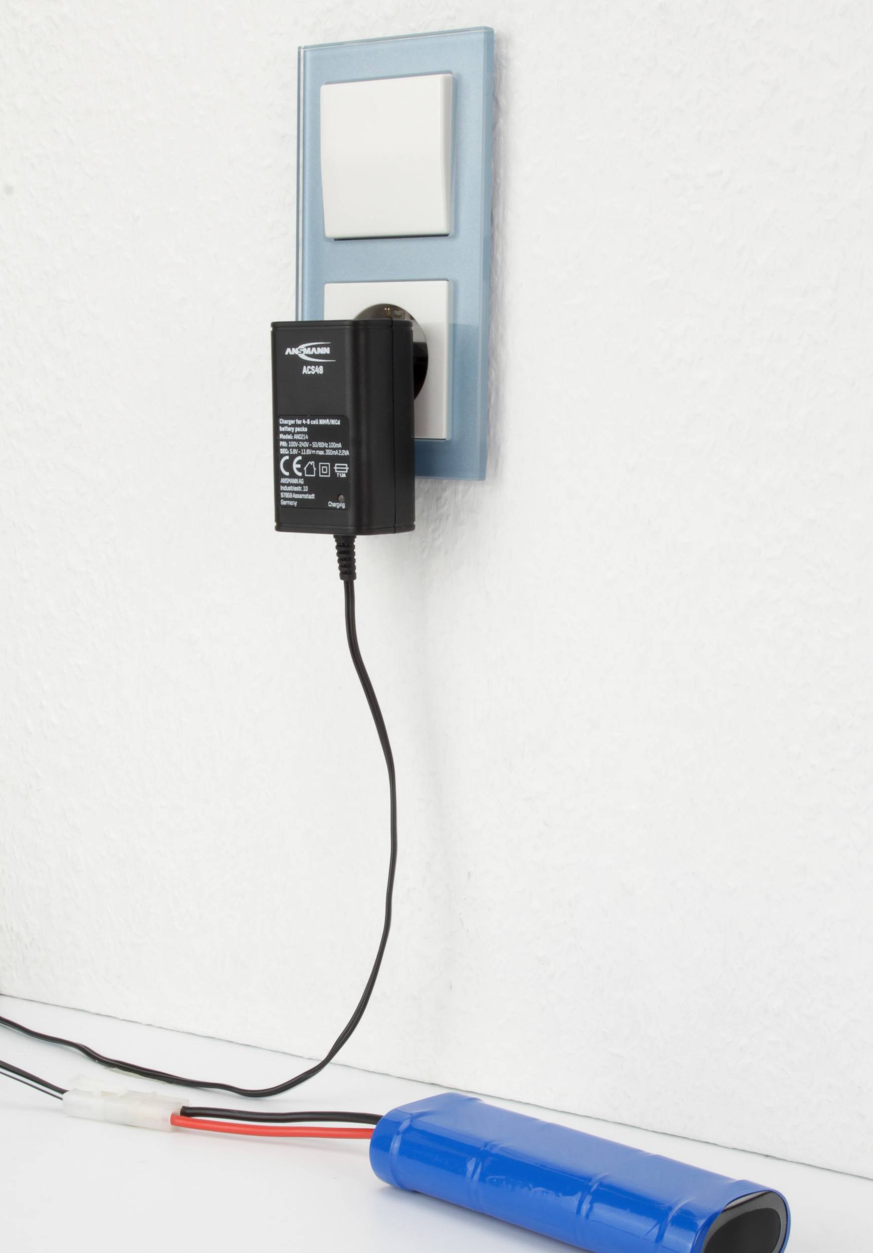 Ein Ladegerät ist in die Steckdose an einer weißen Wand eingesteckt. Ein Kabel führt zu einem blauen Akkupack auf einer weißen Oberfläche.