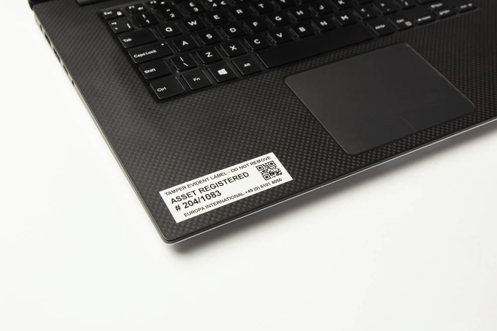 Ein Laptop mit schwarzer Tastatur zeigt ein Etikett mit der Aufschrift 'Asset Registered' und einer Seriennummer auf der Handballenauflage.