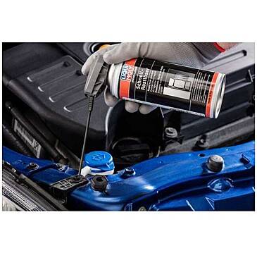 Die behandschuhte Hand einer Person sprüht Schmierspray aus einer Dose auf eine blaue Metallkomponente eines Automotors, um Quietschgeräusche zu vermeiden.