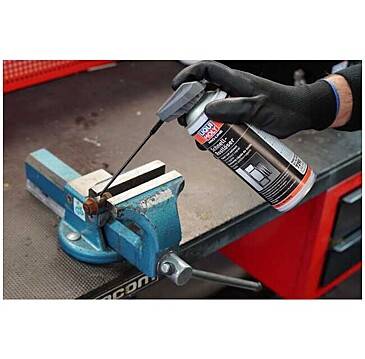 Eine behandschuhte Hand sprüht Schmiermittel auf ein Metallteil, das in einer blauen Klemme auf einer Werkbank gehalten wird.