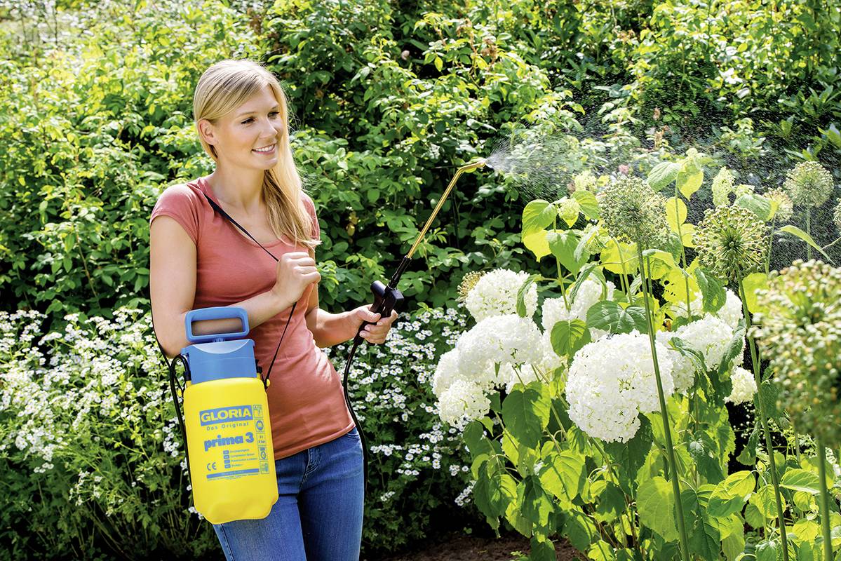 Eine Frau sprüht im Garten Pflanzen mit einem gelben Drucksprüher an. Sie trägt lässige Kleidung und steht vor grünen Sträuchern.