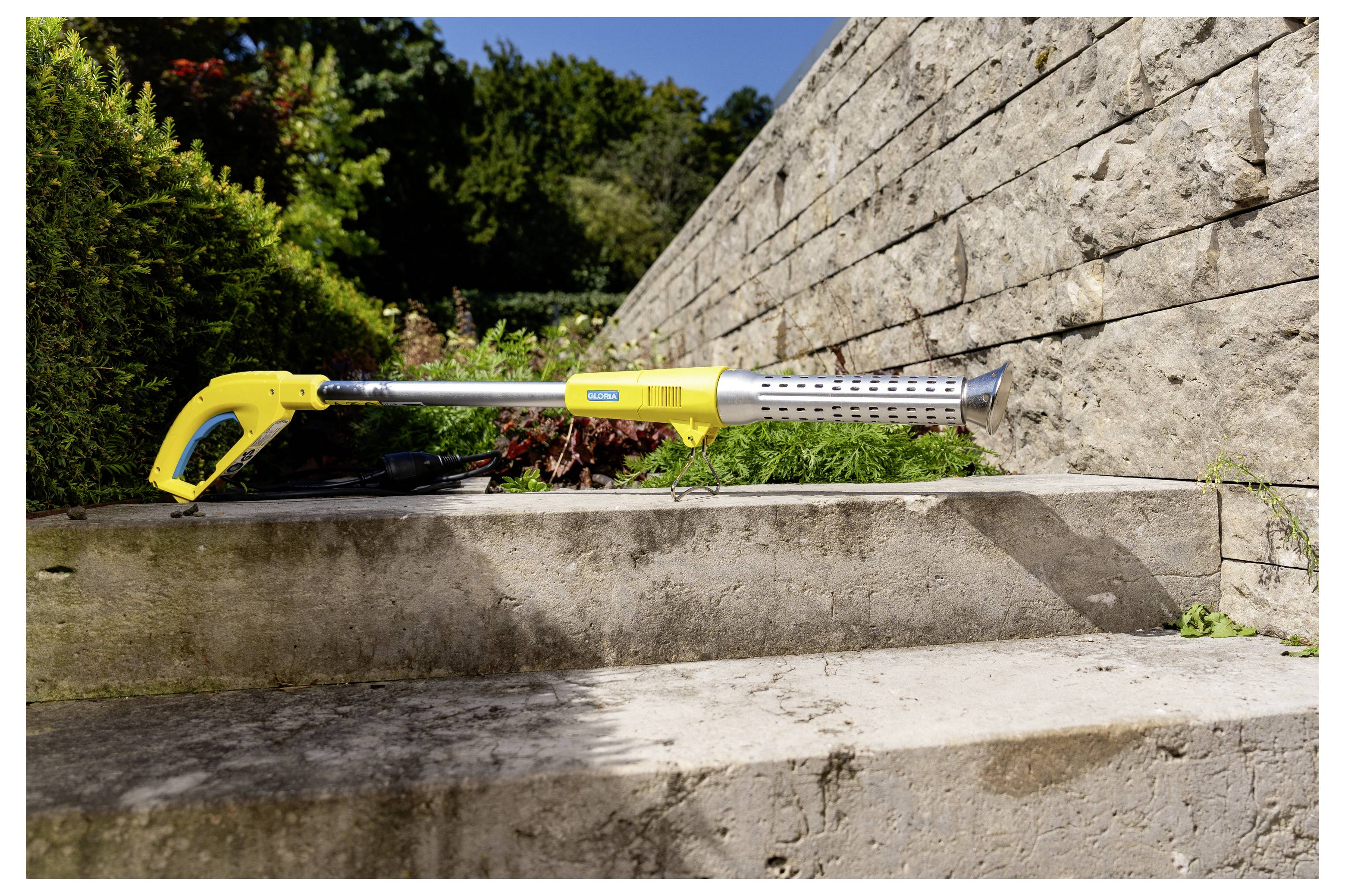 Gelber tragbarer Laubbläser, der auf Betontreppenstufen neben einer Steinmauer im Freien bei hellem Sonnenlicht ruht.