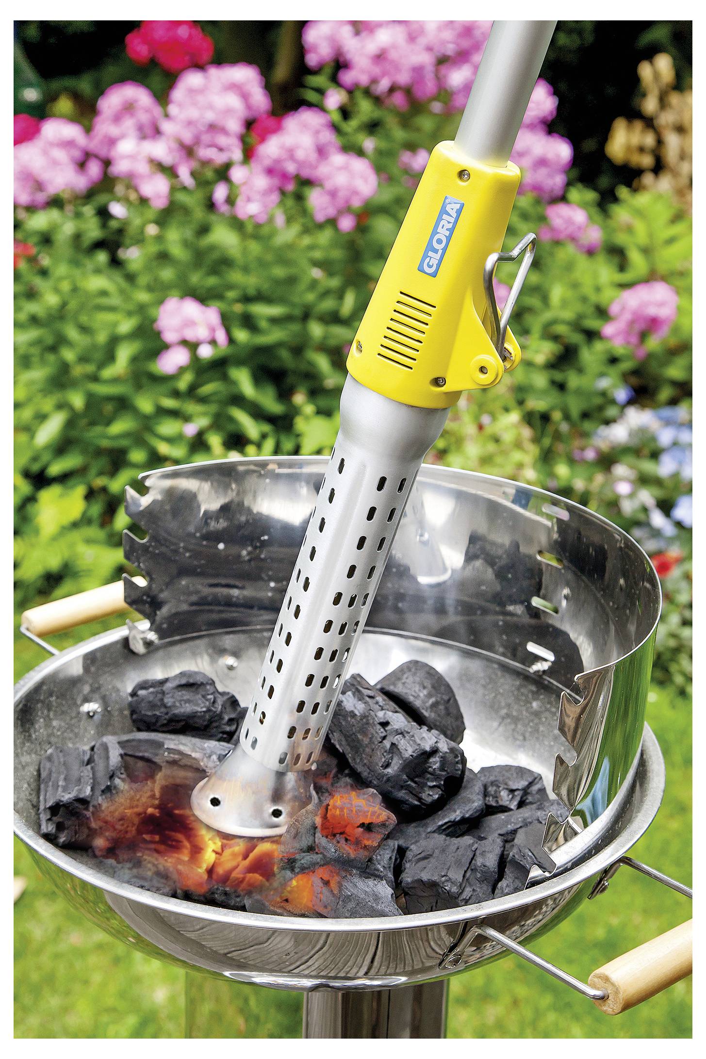 Elektrischer Holzkohlestarter heizt Kohlen in einem runden Outdoor-Grill mit glühenden Glut auf, während Gartenblumen im Hintergrund verschwommen sind.