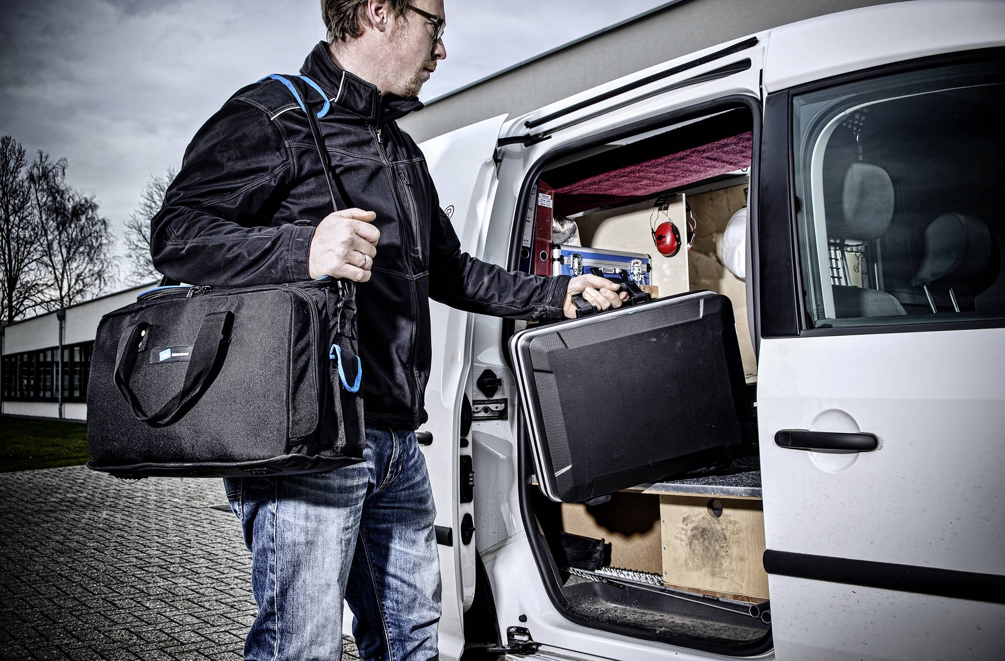Ein Mann lädt Koffer und Taschen mit Ausrüstung in einen weißen Lieferwagen, im Hintergrund sind ein Gebäude und Bäume sichtbar.