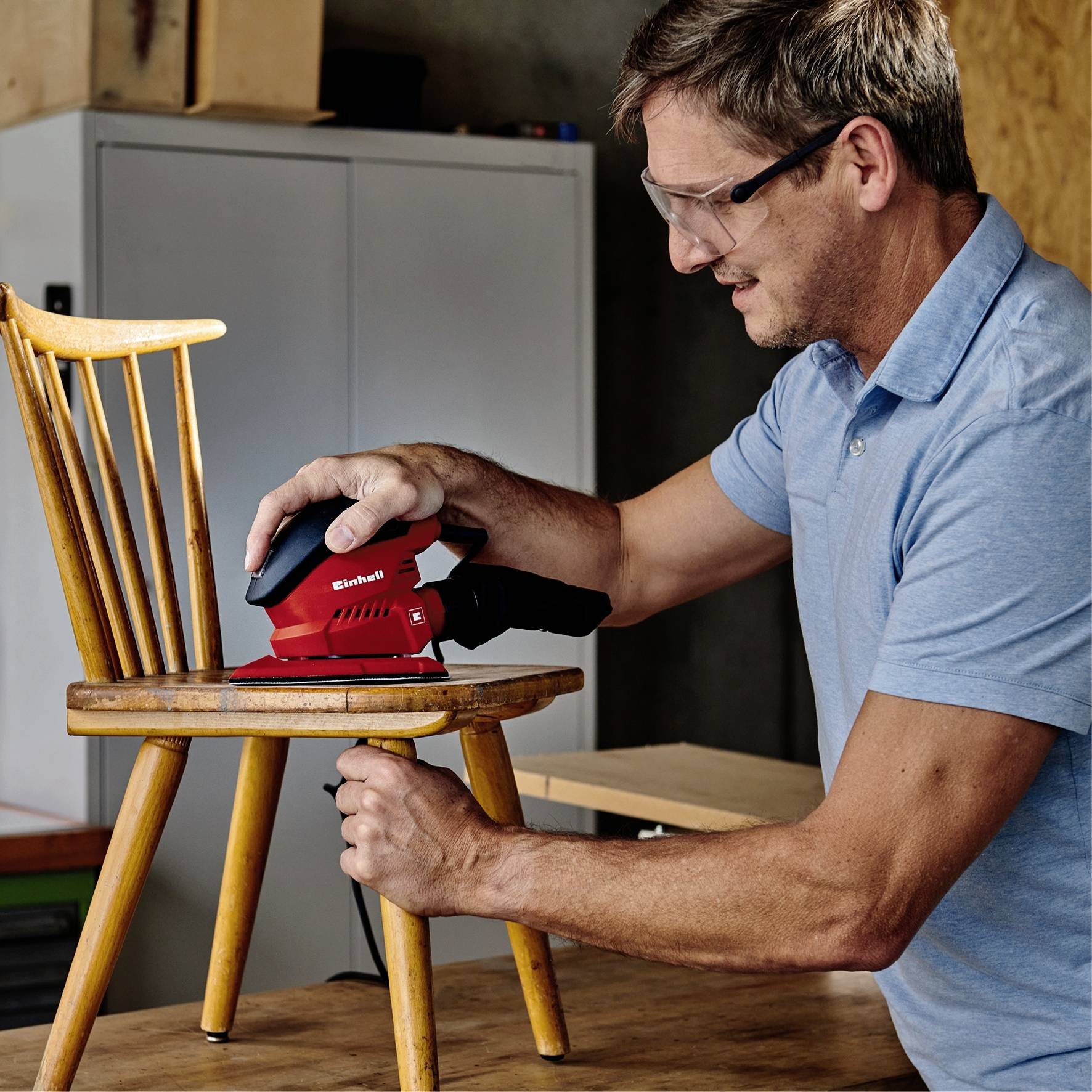 Ein Mann schleift mit einer elektrischen Schleifmaschine einen Holzstuhl. Er trägt eine Schutzbrille und arbeitet konzentriert.