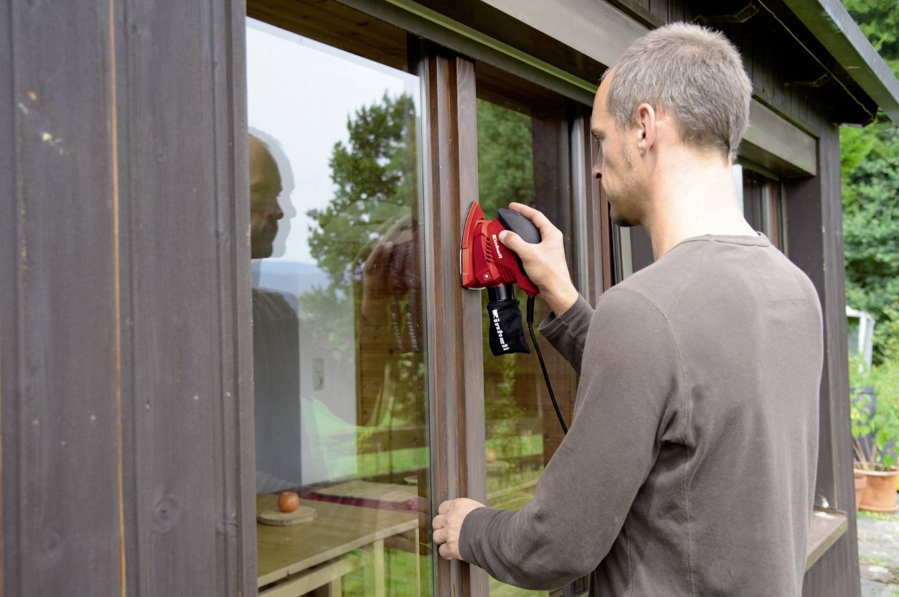 Ein Mann reinigt mit einem roten Gerät ein Fenster von außen. Der Hintergrund zeigt Bäume und einen Garten.