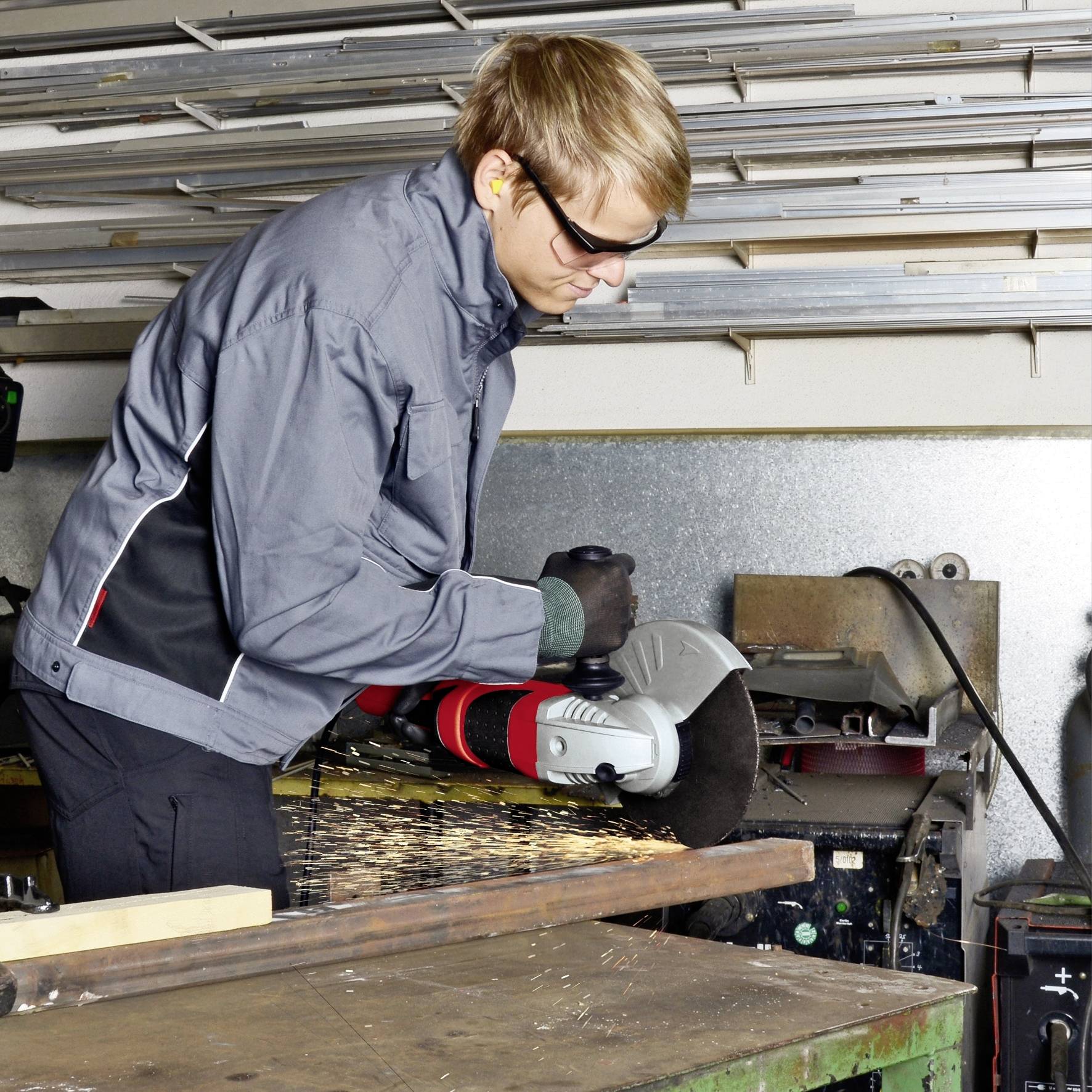 Eine Person in Schutzkleidung schneidet mit einem Winkelschleifer Metall in einer Werkstatt. Funken fliegen vom Werkstück.