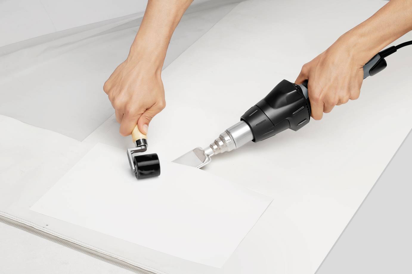 Zwei Hände bei der Verwendung eines Heißluftföns und einer Rolle, um Kunststoffmaterial zu verschweißen.