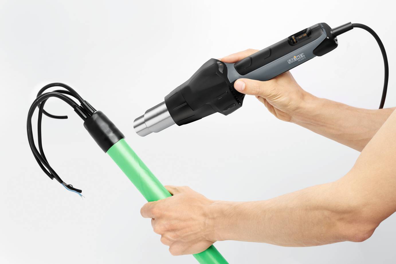 Eine Person hält ein Heißluftgebläse und erwärmt damit das Ende eines grünen Rohrs, aus dem schwarze Kabel hervorschauen.