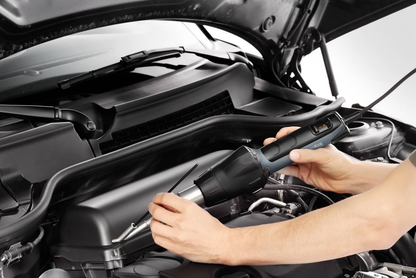 Eine Person repariert einen Auto-Motor mit einem elektrischen Werkzeug. Das Bild zeigt detailliert die Motorhaube und das Werkzeug.