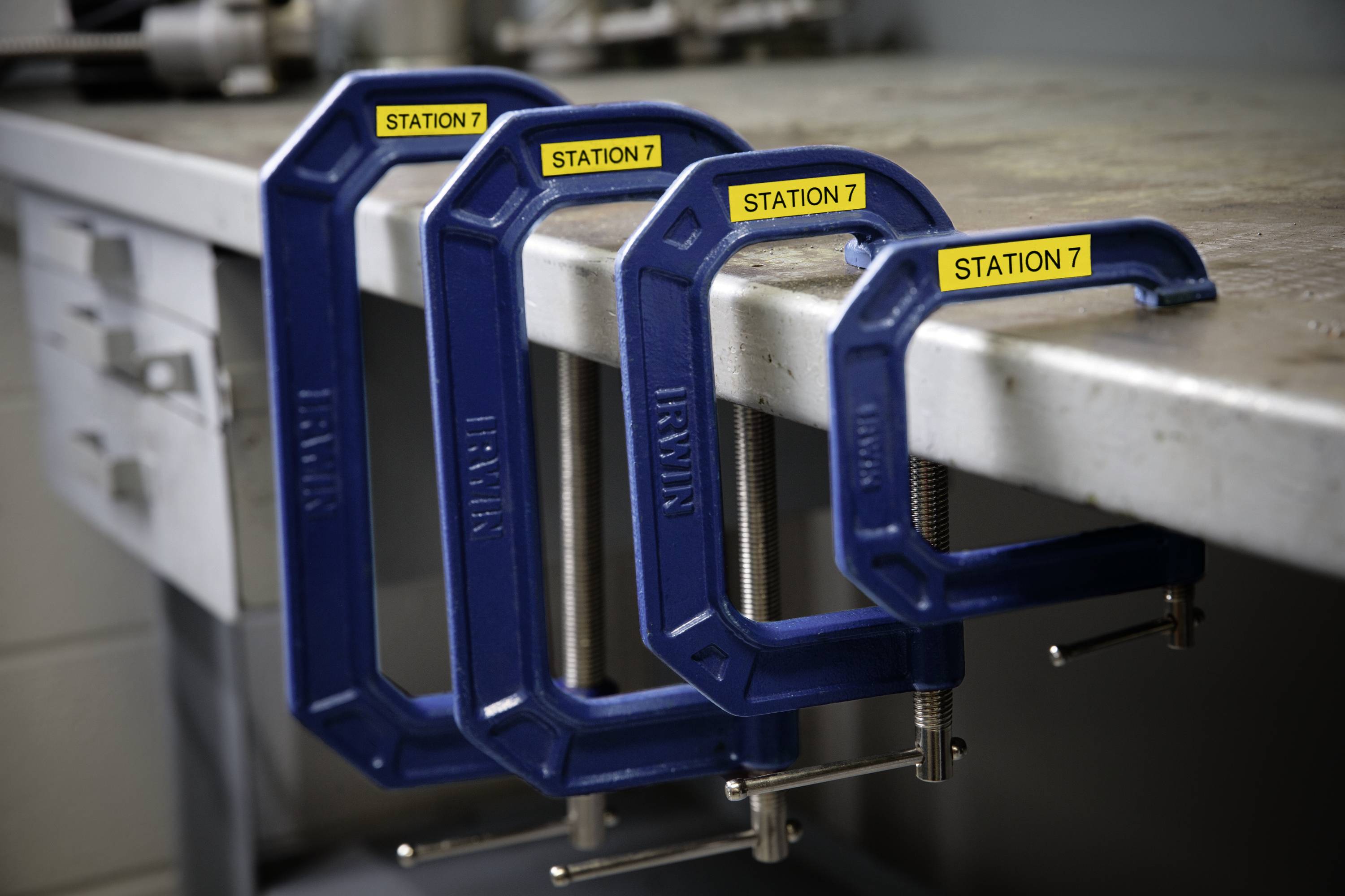 Vier blaue Schraubzwingen unterschiedlicher Größe klemmen an einer Werkbankkante. Jede Zwinge ist mit 'Station 7' beschriftet.