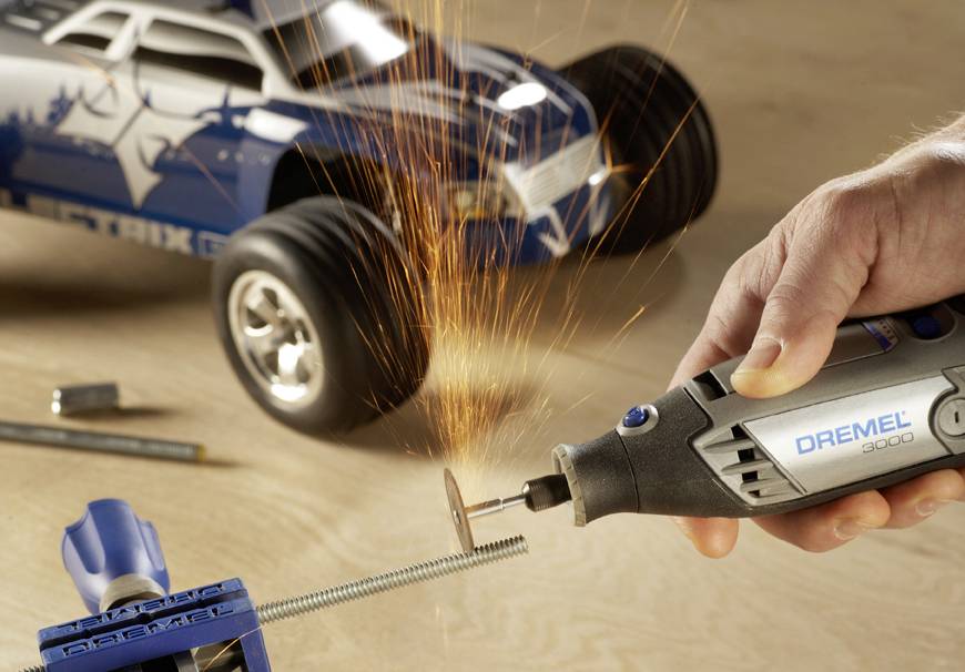 Person schneidet eine Schraube mit einem Dremel-Werkzeug, während im Hintergrund ein Modellauto steht. Funken sprühen beim Schneiden.