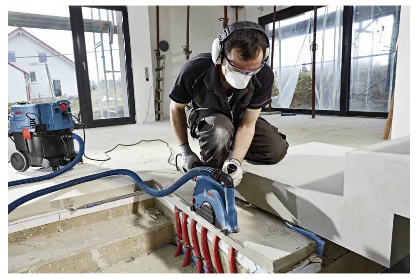 Ein Arbeiter nutzt ein Elektrowerkzeug, um Beton in einem teilweise konstruierten Gebäude zu schneiden, trägt Schutzausrüstung und kniet neben Treppen.