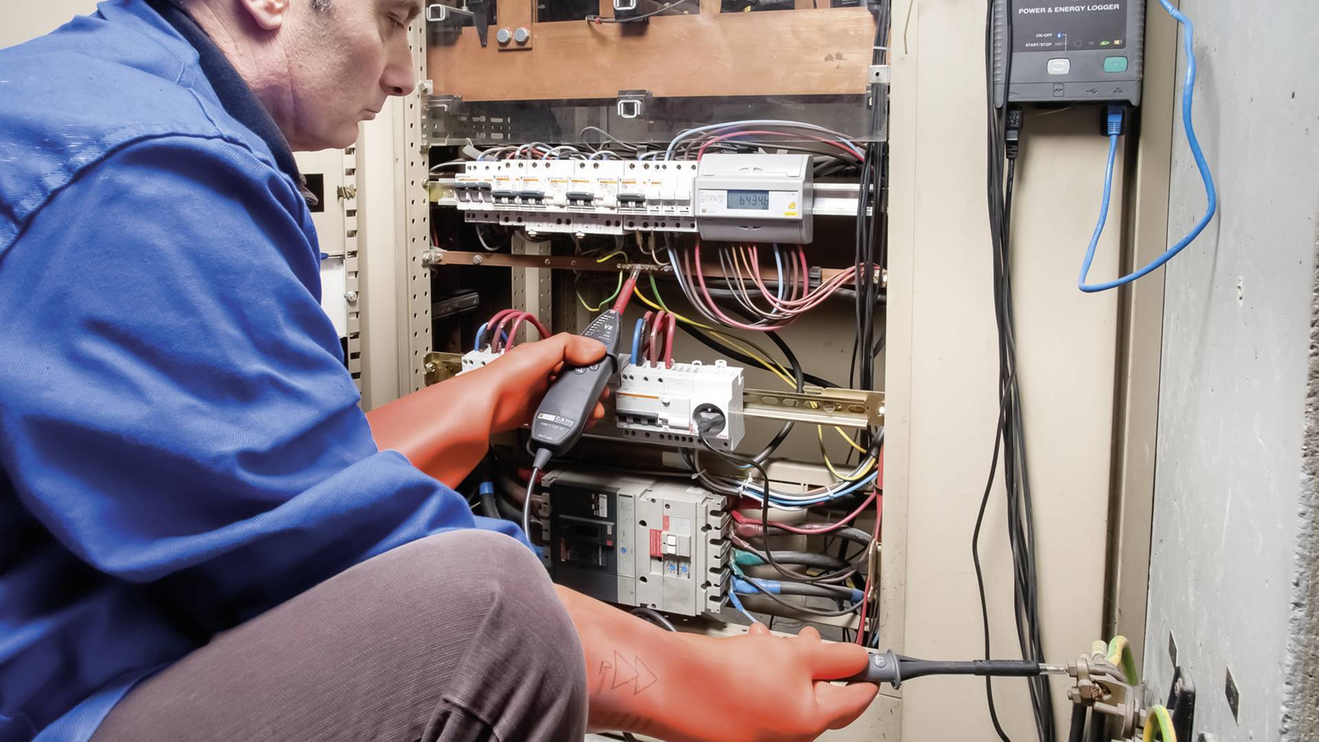 Eine Person in blauer Arbeitskleidung prüft elektrische Schaltungen in einem offenen Schaltschrank mit Kabeln und Geräten.