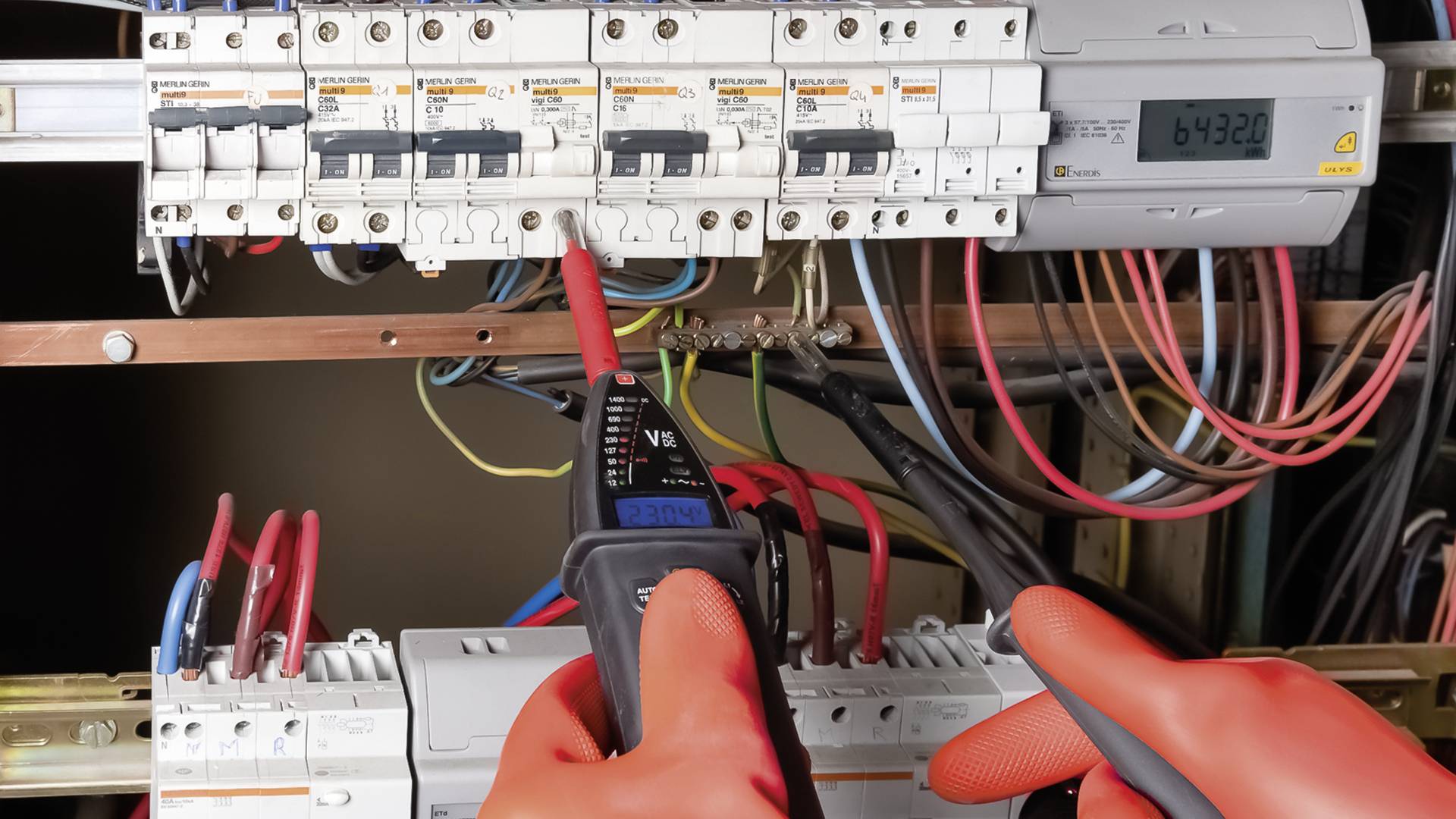 Person mit roten Schutzhandschuhen prüft elektrische Schaltkreise in einem Sicherungskasten mit einem Multimeter.