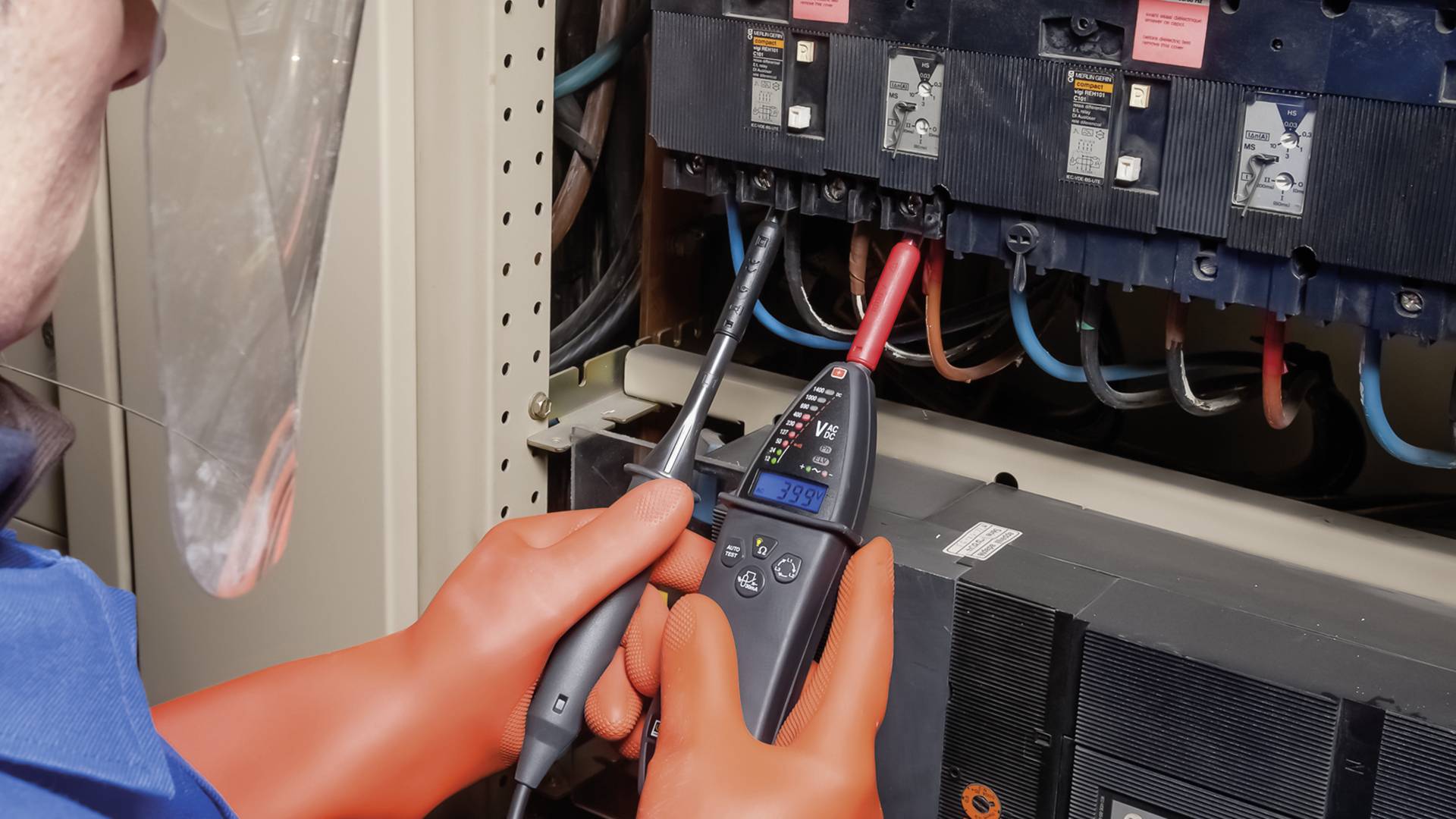 Eine Person mit Schutzbrille und -handschuhen misst Spannung an einem offenen Sicherungskasten mit einem digitalen Multimeter.