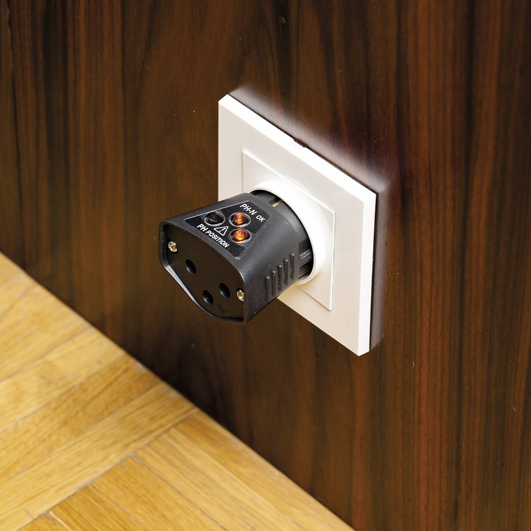 Ein Stromprüfgerät steckt in der Steckdose einer Holzverkleidung. Es zeigt an, ob die elektrische Verbindung sicher und korrekt ist.