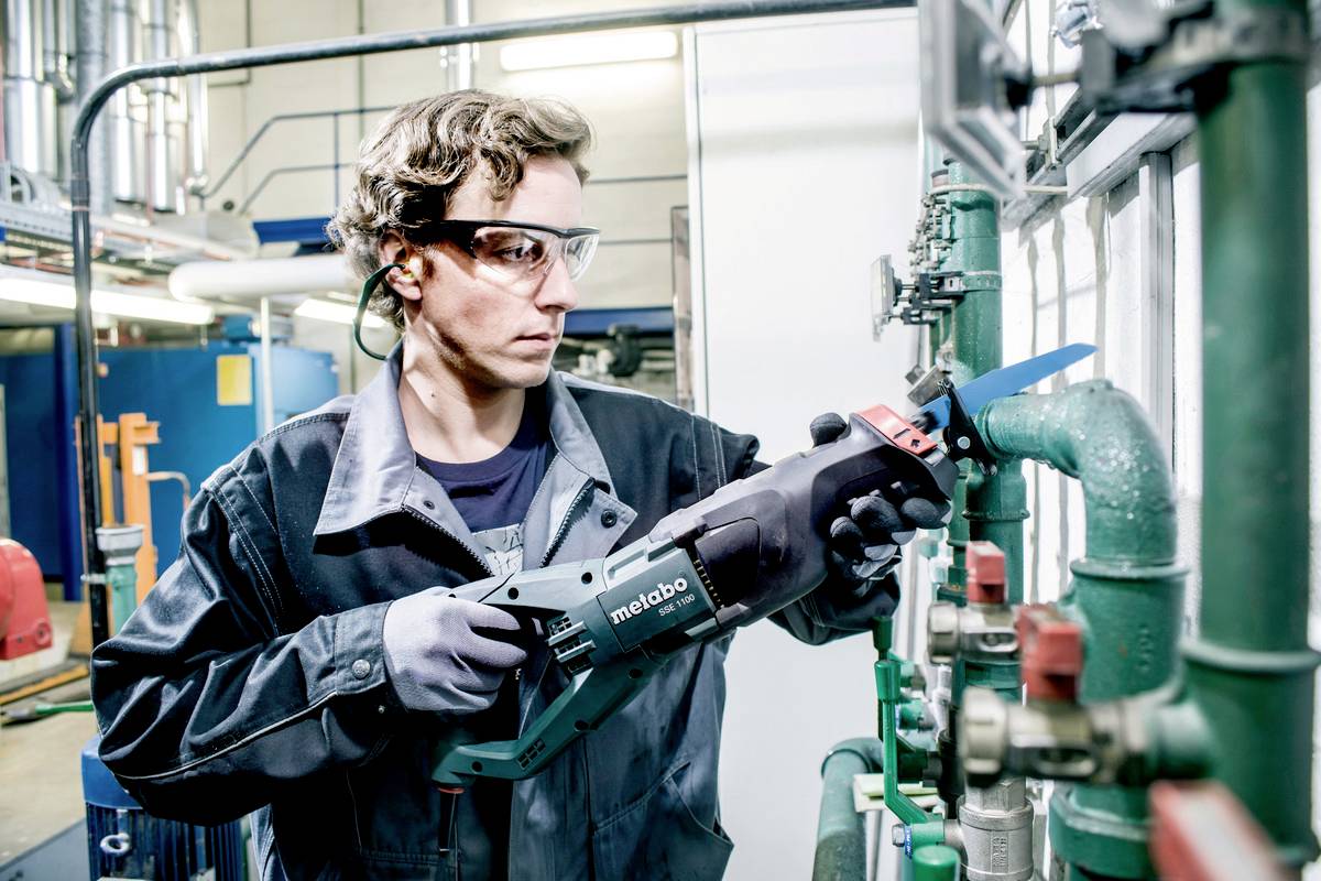 Ein Arbeiter in Schutzbrille und Handschuhen verwendet eine Metabo-Säbelsäge, um an Rohren in einer industriellen Umgebung zu arbeiten.