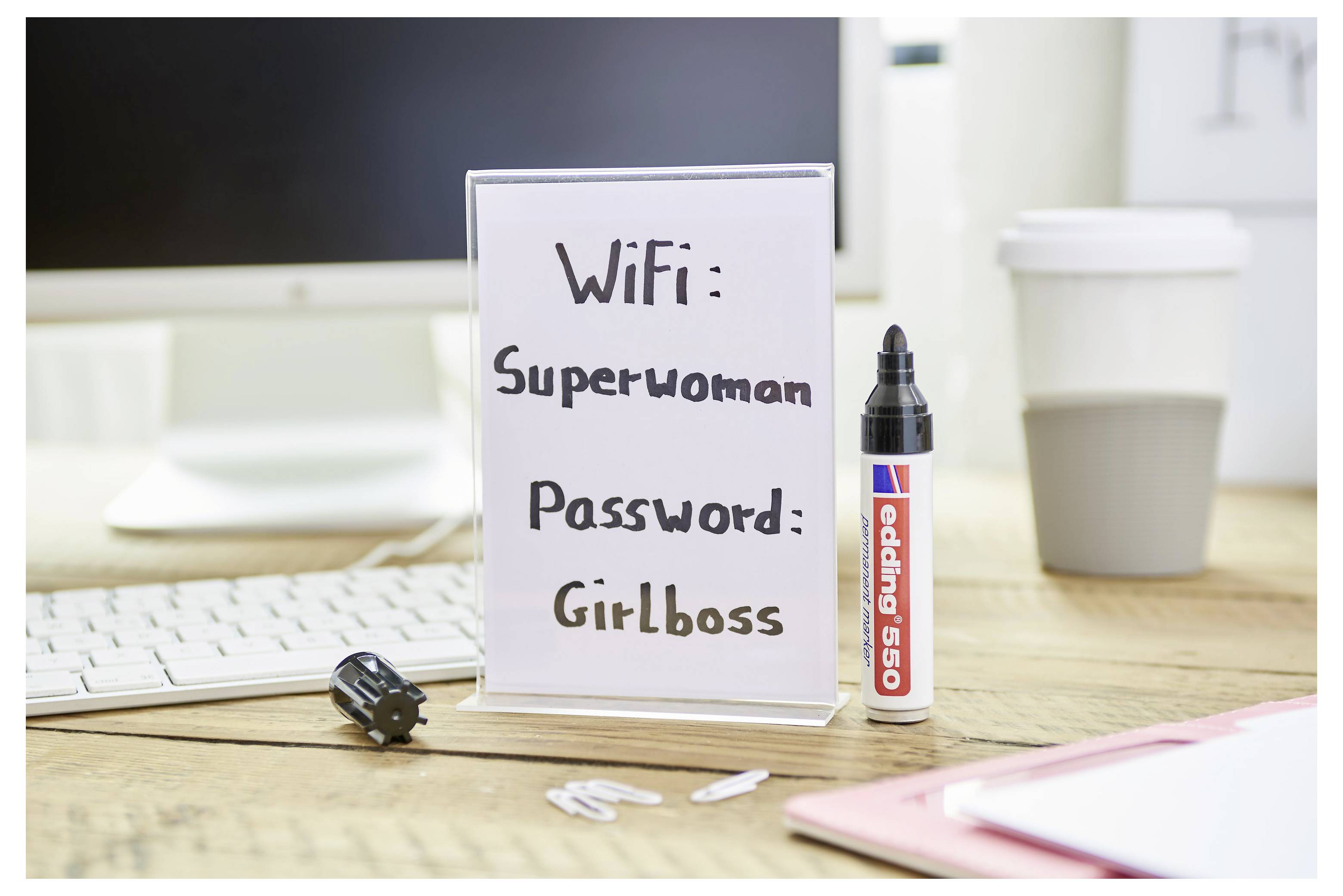 Schreibtischschild zeigt WLAN-Anmeldedaten: Benutzername „Superwoman" und Passwort „Girlboss", neben einem Marker, einer Tastatur und einer Kaffeetasse.