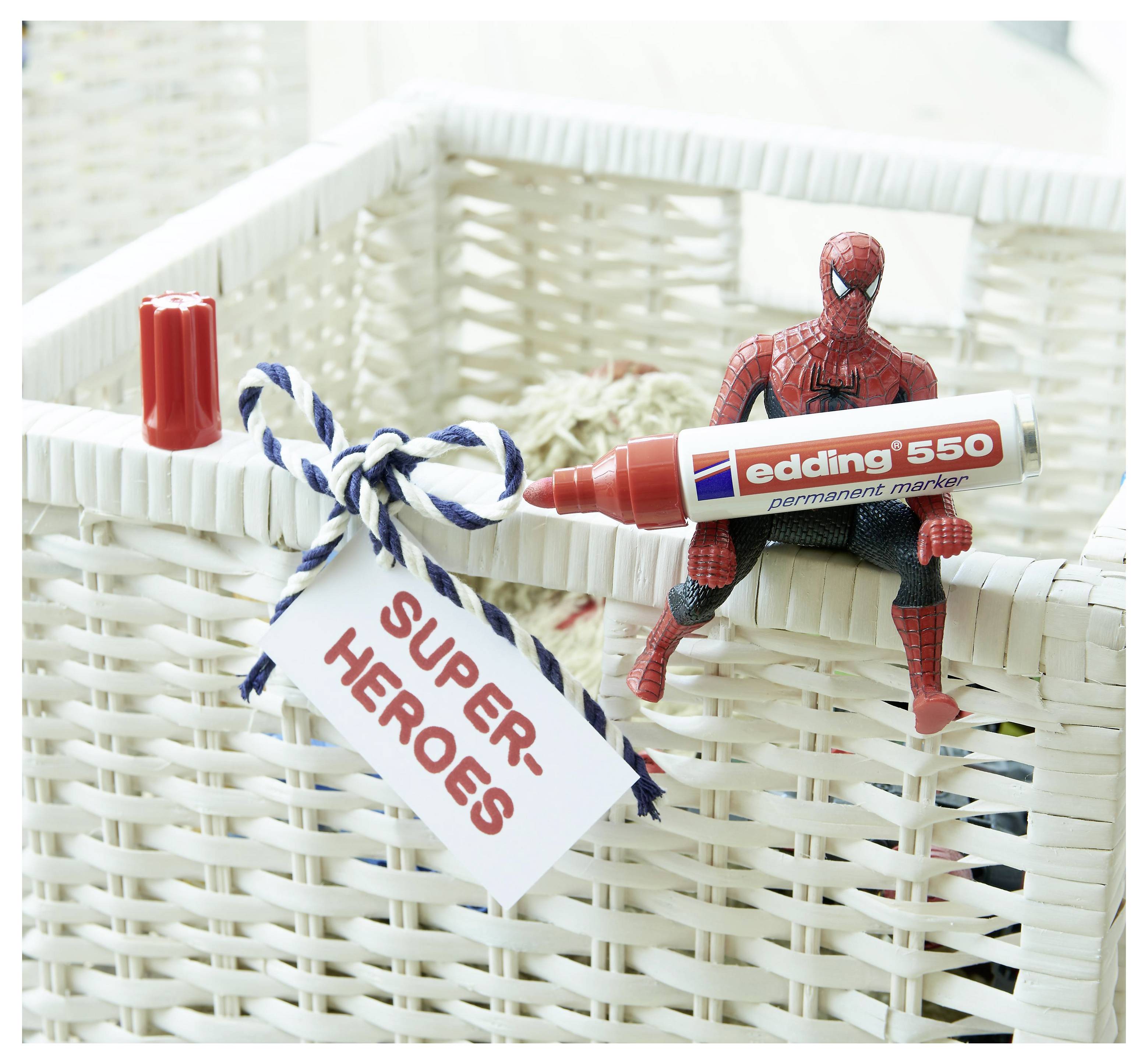 Spider-Man-Figur sitzt auf einem weißen Korbgeflecht und hält einen roten „edding 550"-Marker, mit einem Hängeschild, das „SUPER HEROES" liest.