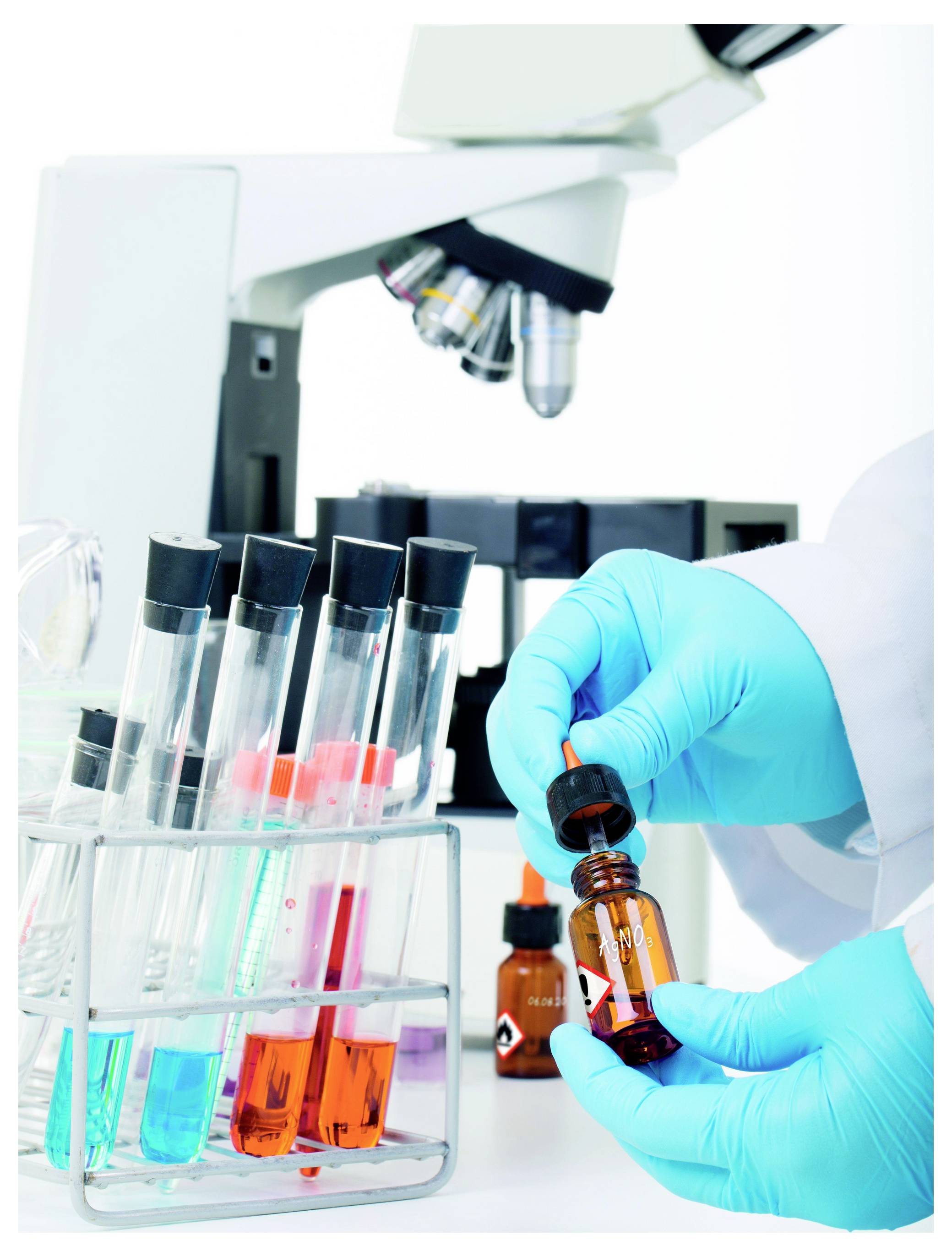 Laborant mit Handschuhen, der eine Tropferflasche neben Reagenzgläsern und einem Mikroskop in einem wissenschaftlichen Labor handhabt.