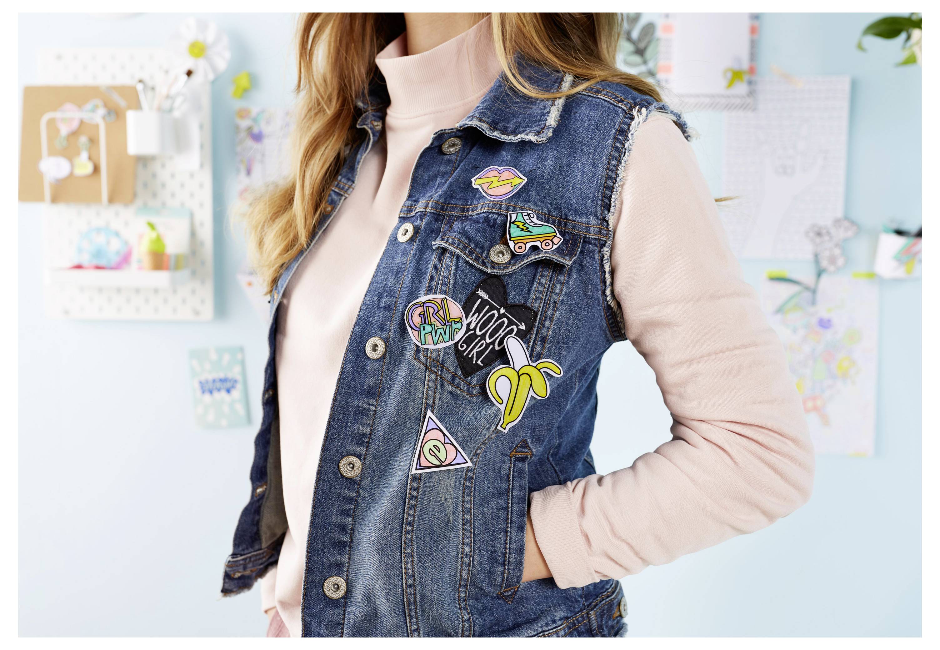 Frau, die eine Jeansweste mit farbenfrohen bestickten Aufnähern über einem rosa Top trägt, in einem hellen Raum mit Wandkunst im Hintergrund dargestellt.