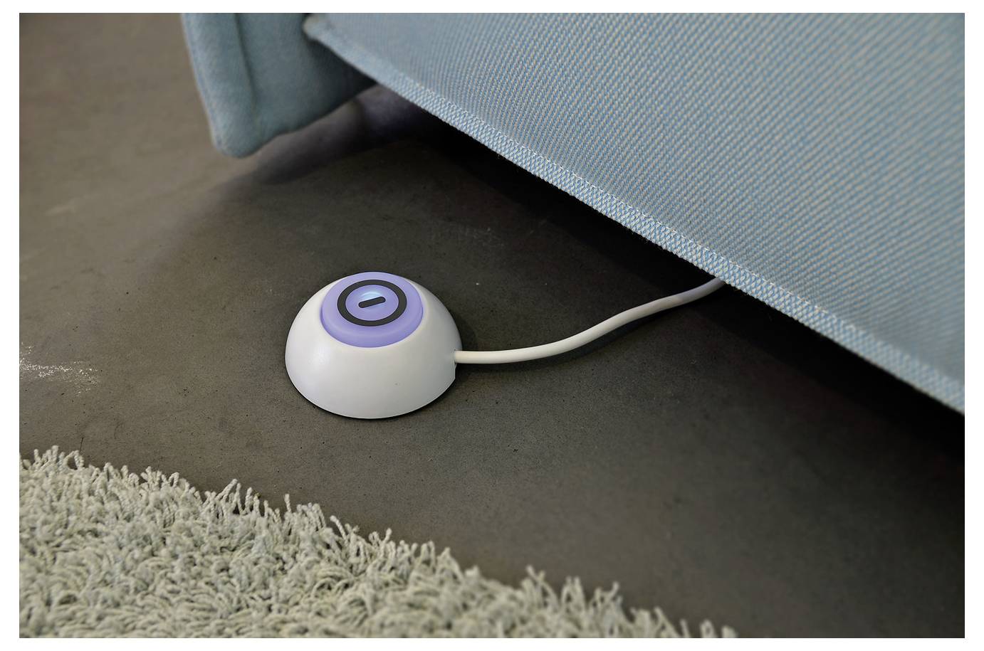 Kleiner runder weißer Bodenknopf mit blauem Stromsymbol neben einem Sofa, wahrscheinlich eine verborgene Steuerung oder ein Bodenschalter.